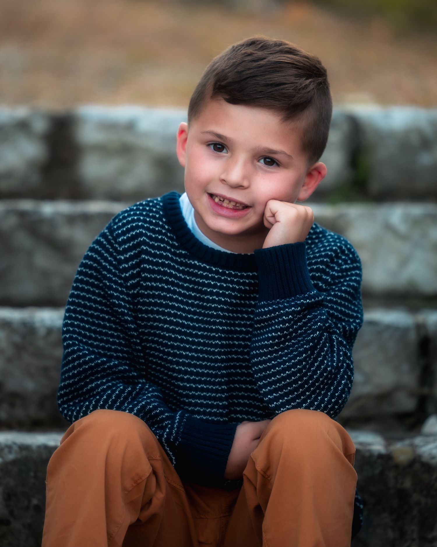 A young child in a navy striped sweater and brown pants sits casually on stone steps outdoors.