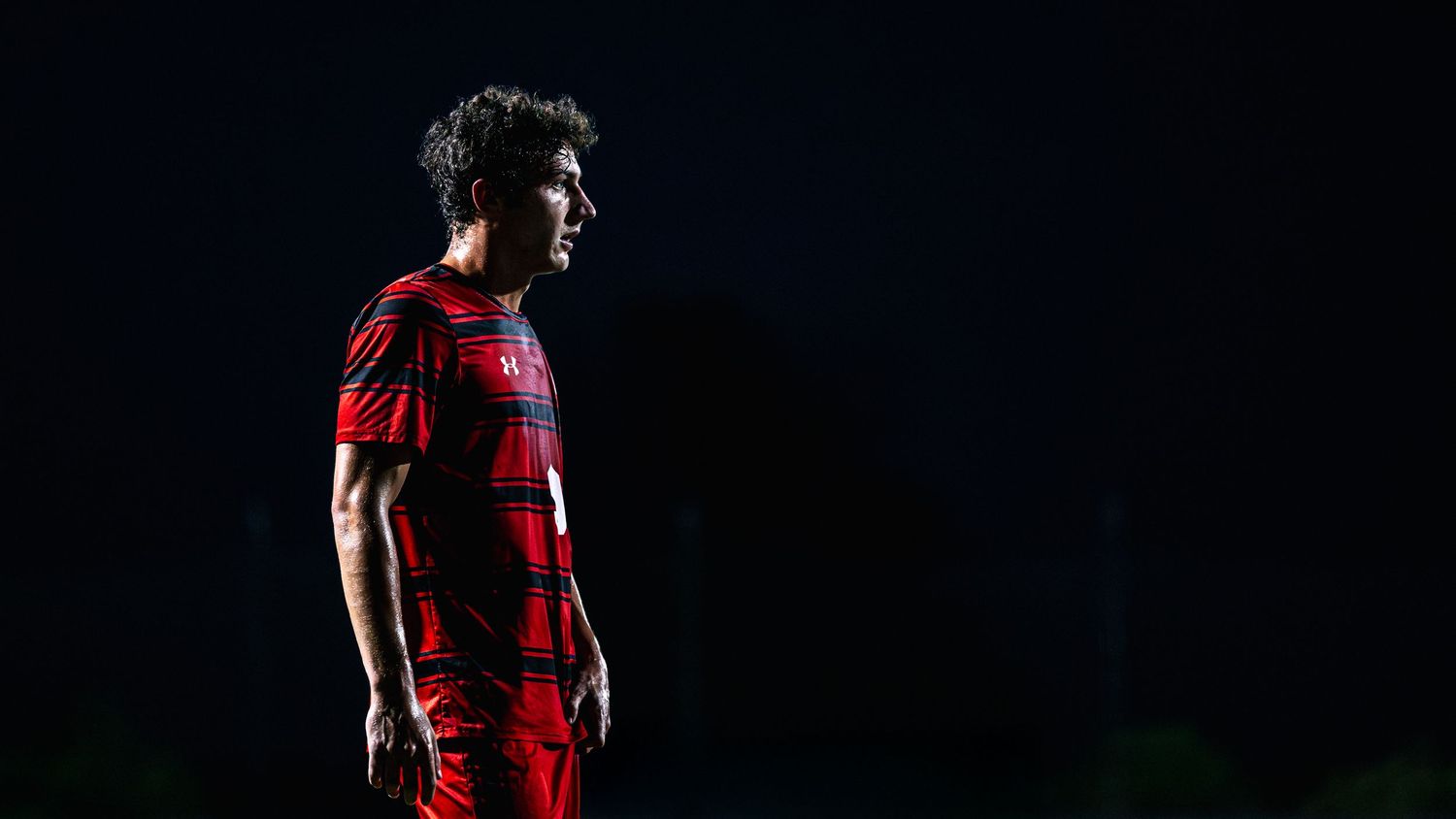 Soccer player in red and black striped uniform running on field during nighttime match.