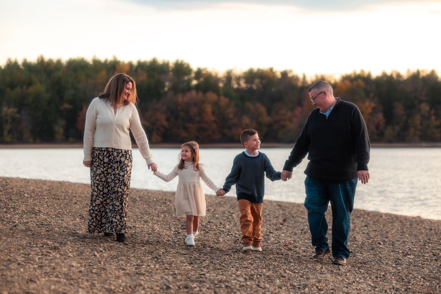 Family walking together along a lakeside beach at sunset with autumn trees in the background.