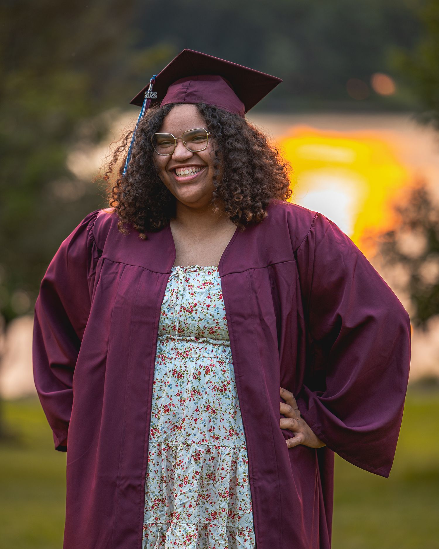 Graduate in maroon cap and gown with floral dress smiles joyfully during outdoor graduation photo session at sunset.