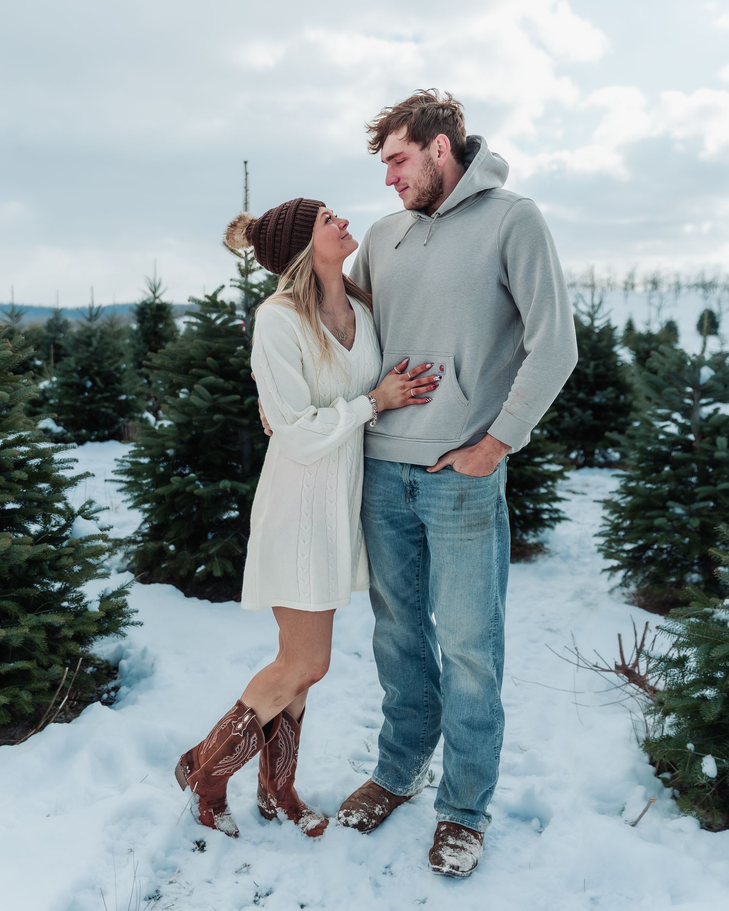 A couple shares romantic moments in a snowy Christmas tree farm during winter, captured in multiple candid poses.