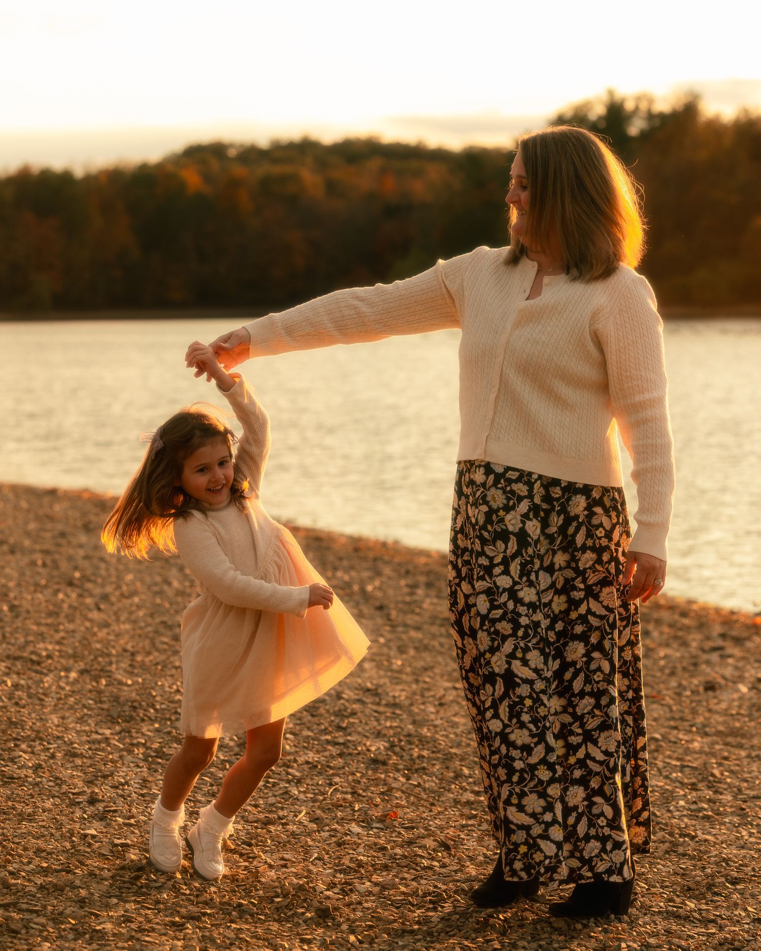 A person in a white dress and another in a floral skirt dance together on a rocky beach at sunset.