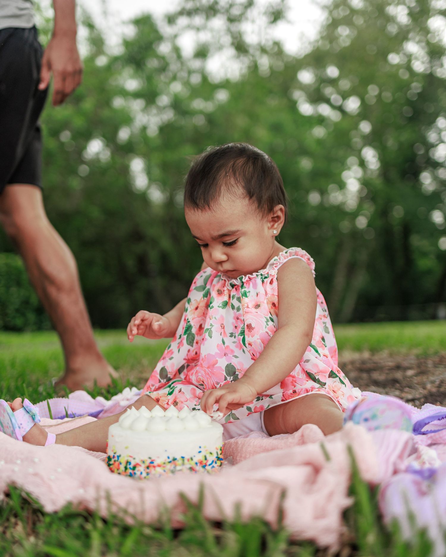 A baby in a floral dress sits on a pink blanket outdoors playing with paper and writing materials.