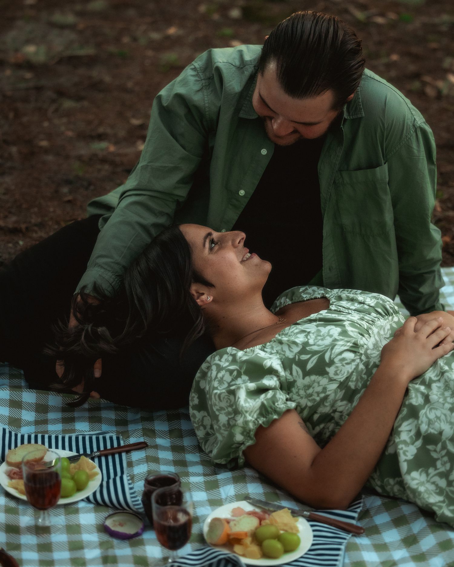 A couple sharing an intimate moment on a picnic blanket with food and drinks laid out on a checkered cloth.