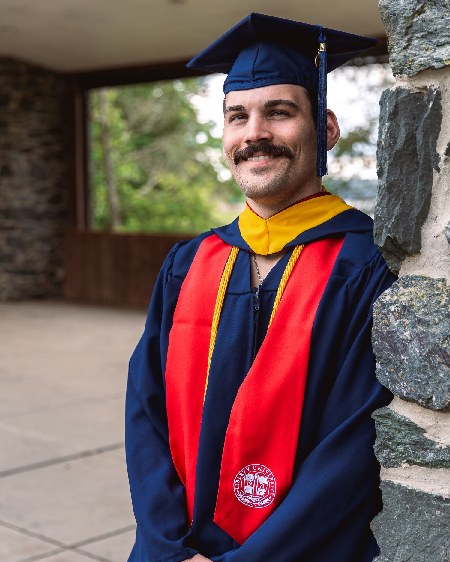 Graduate wearing navy blue and red academic regalia with a graduation cap stands against stone wall backdrop.