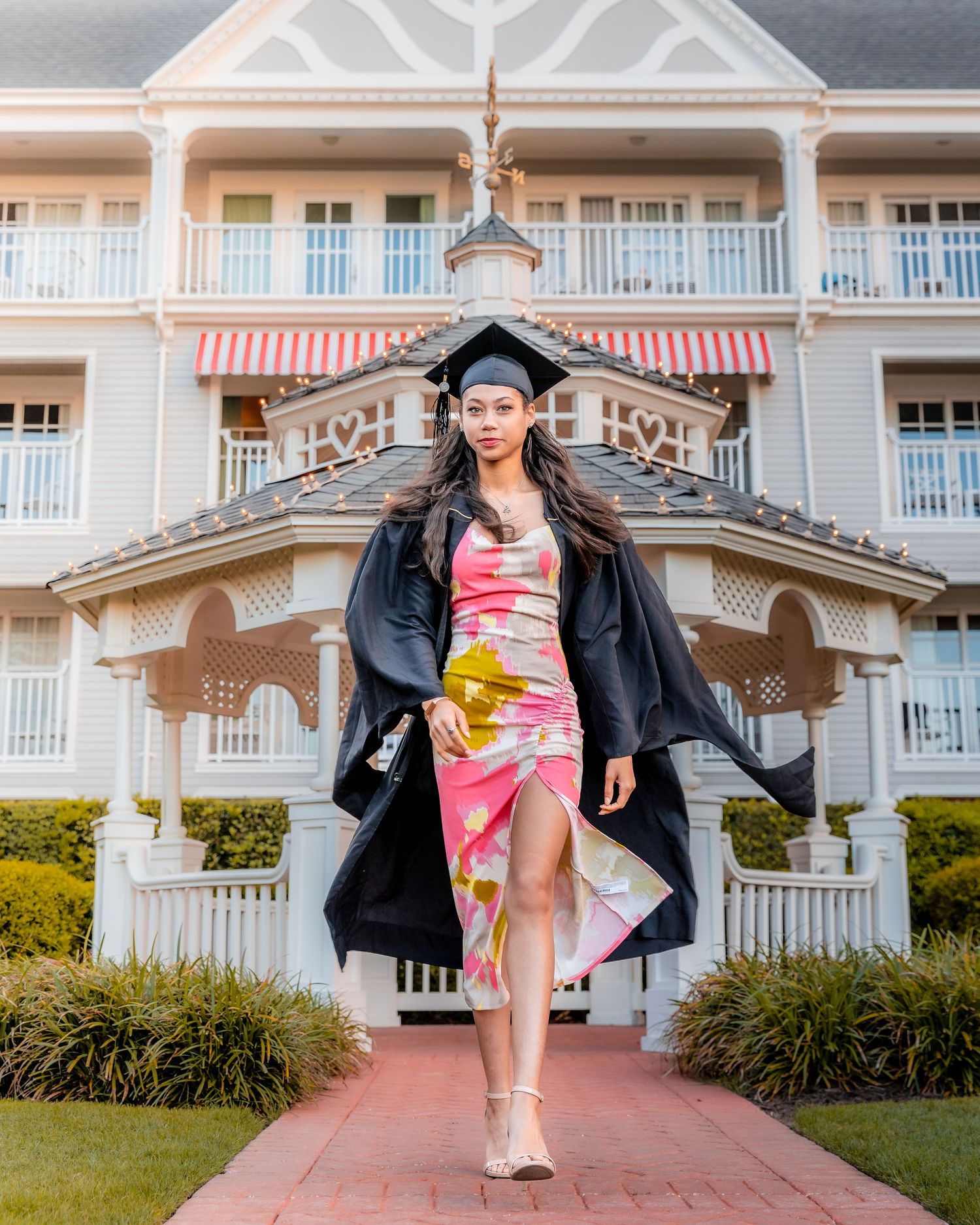 Woman in pink floral dress and black jacket poses on brick path near elegant white building columns.