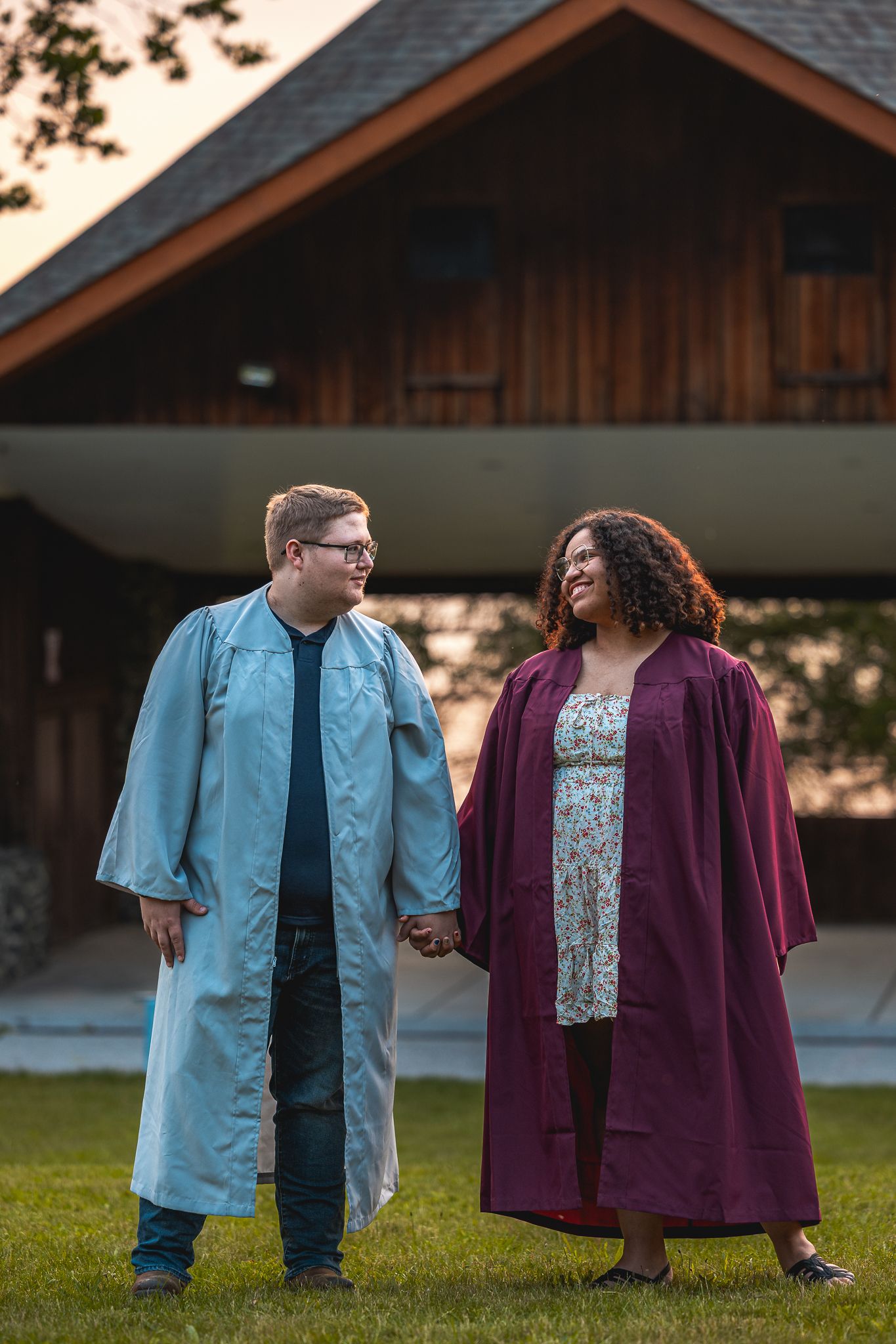 A couple in blue and purple outfits holds hands while walking on grass at sunset.