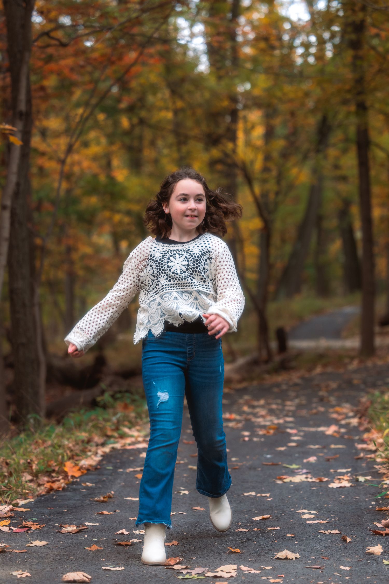 Person in white sweater and blue jeans jumps playfully on autumn path covered with fallen leaves in forest.