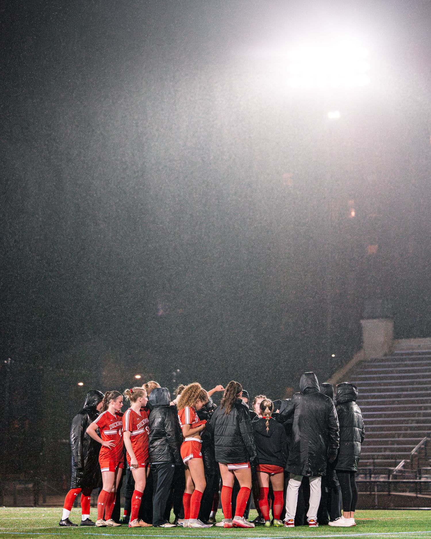 Soccer team huddles together on a floodlit field at night during a match, wearing red uniforms and dark jackets.