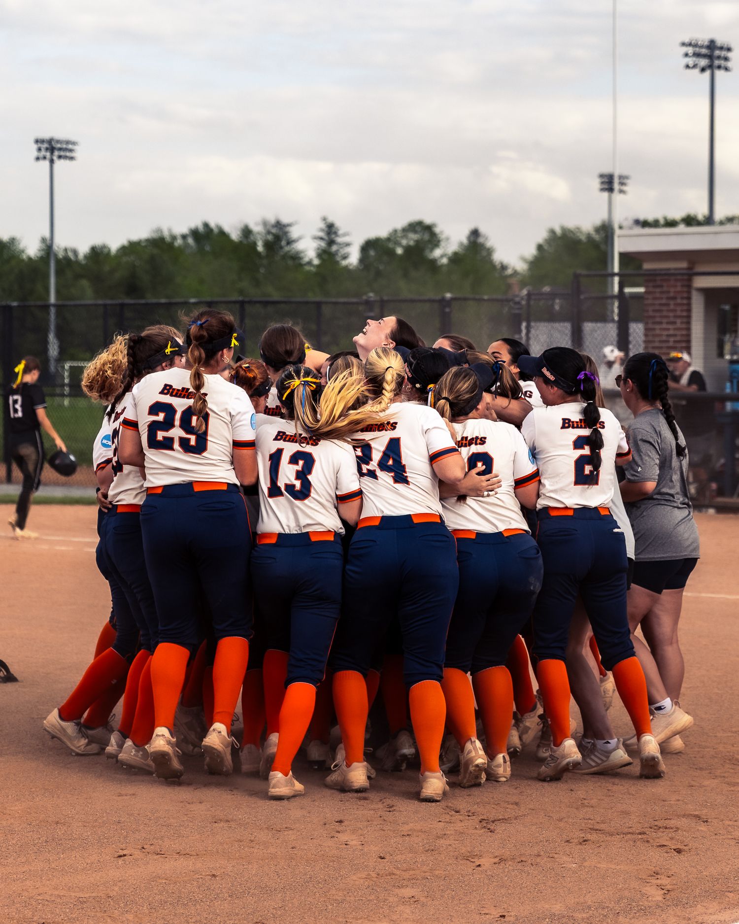 A softball team huddles together in a tight circle on a dirt field during a game or practice session.