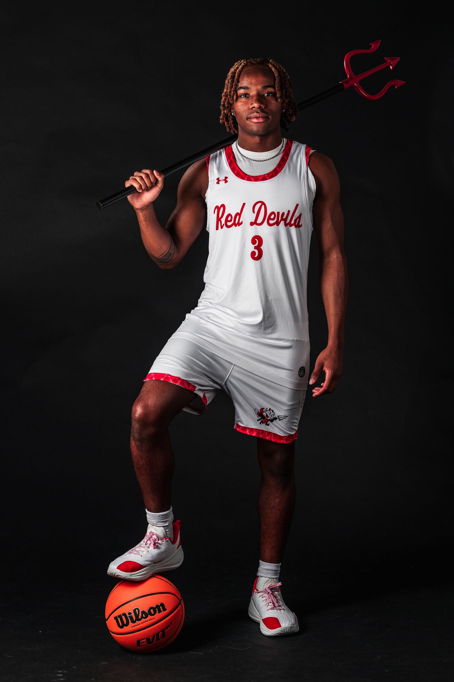 Basketball player in white Red Devils uniform poses with Wilson basketball against dark background in dramatic lighting.