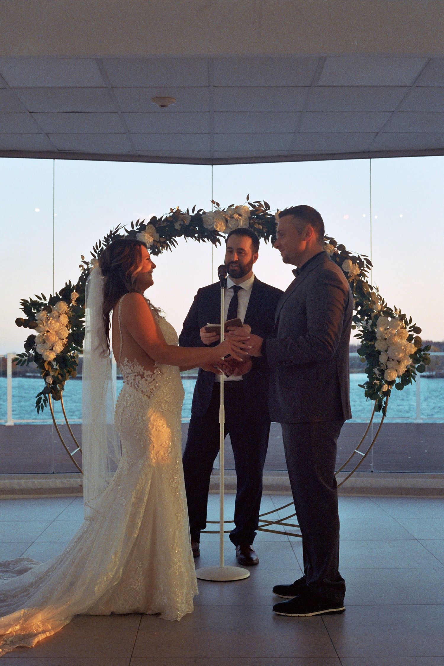 A wedding ceremony takes place under a floral arch with ocean views through large windows in the background.