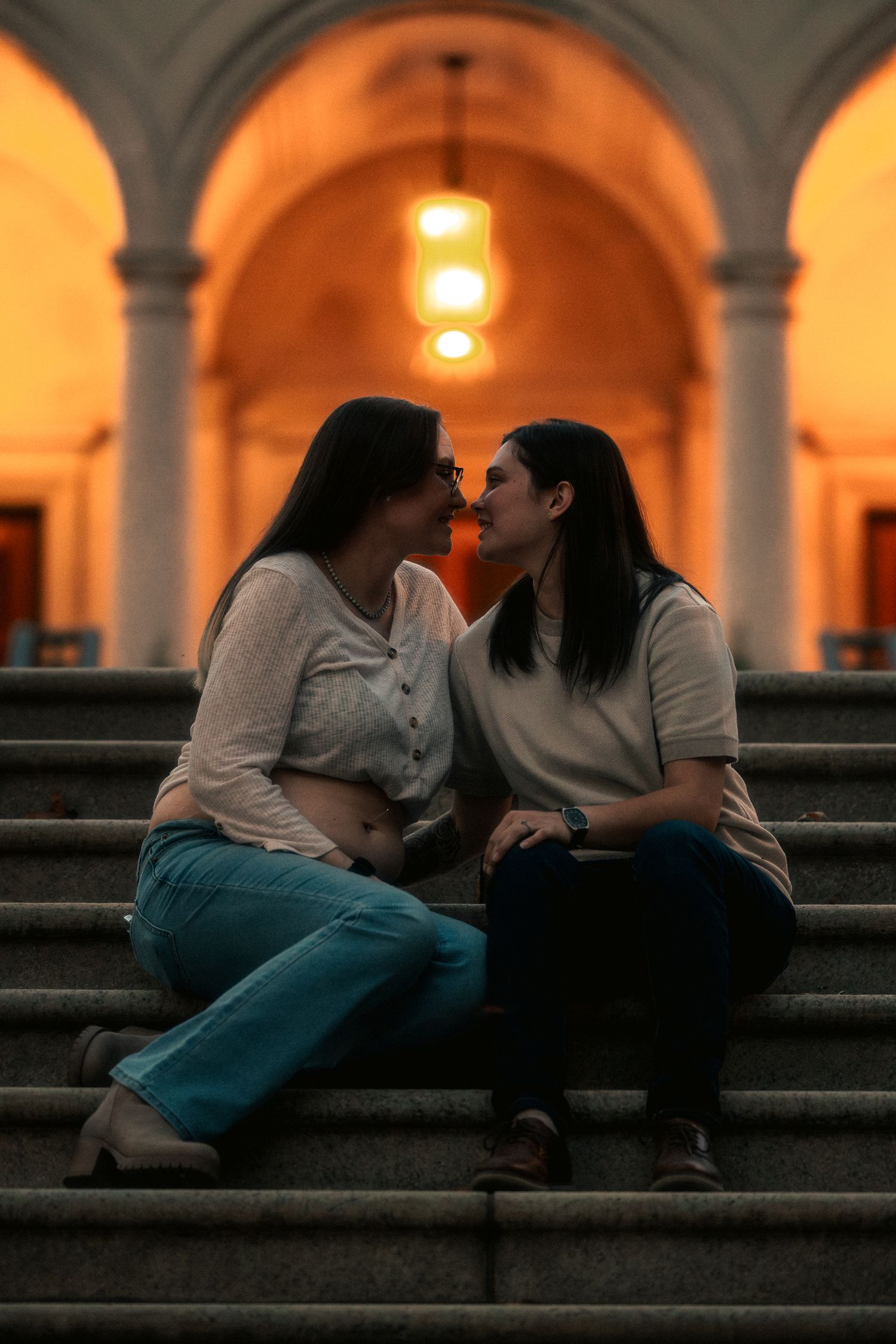 Two people share an intimate moment sitting on stone steps at sunset beneath arched columns with glowing lights.