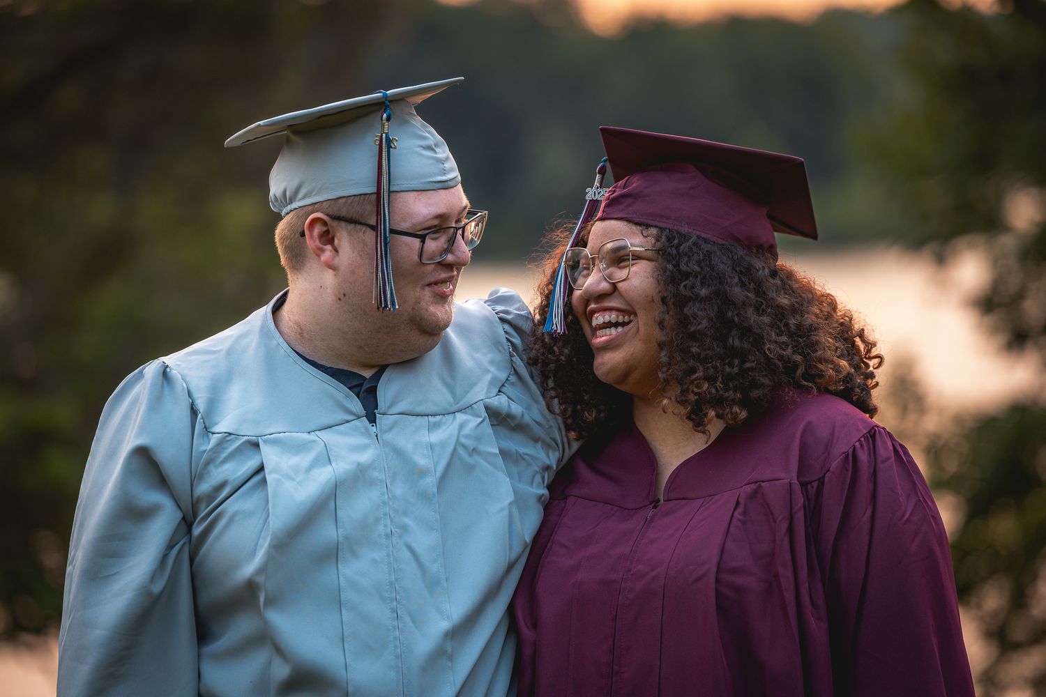 Two graduates in caps and gowns, one in light blue and one in maroon, share a joyful moment during commencement.