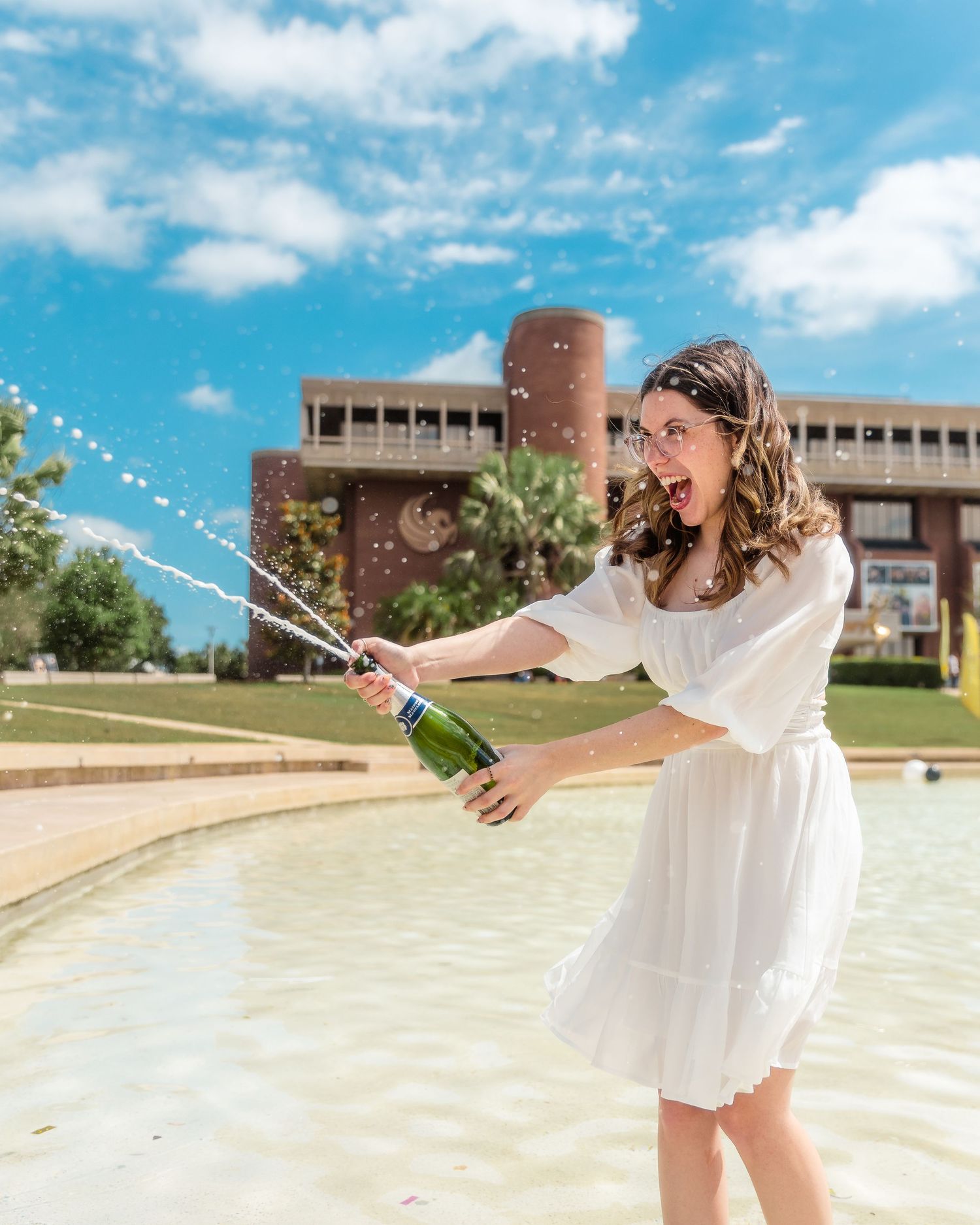 A person in a white dress spins and sprays champagne in celebration on a sunny day outside a brick building.