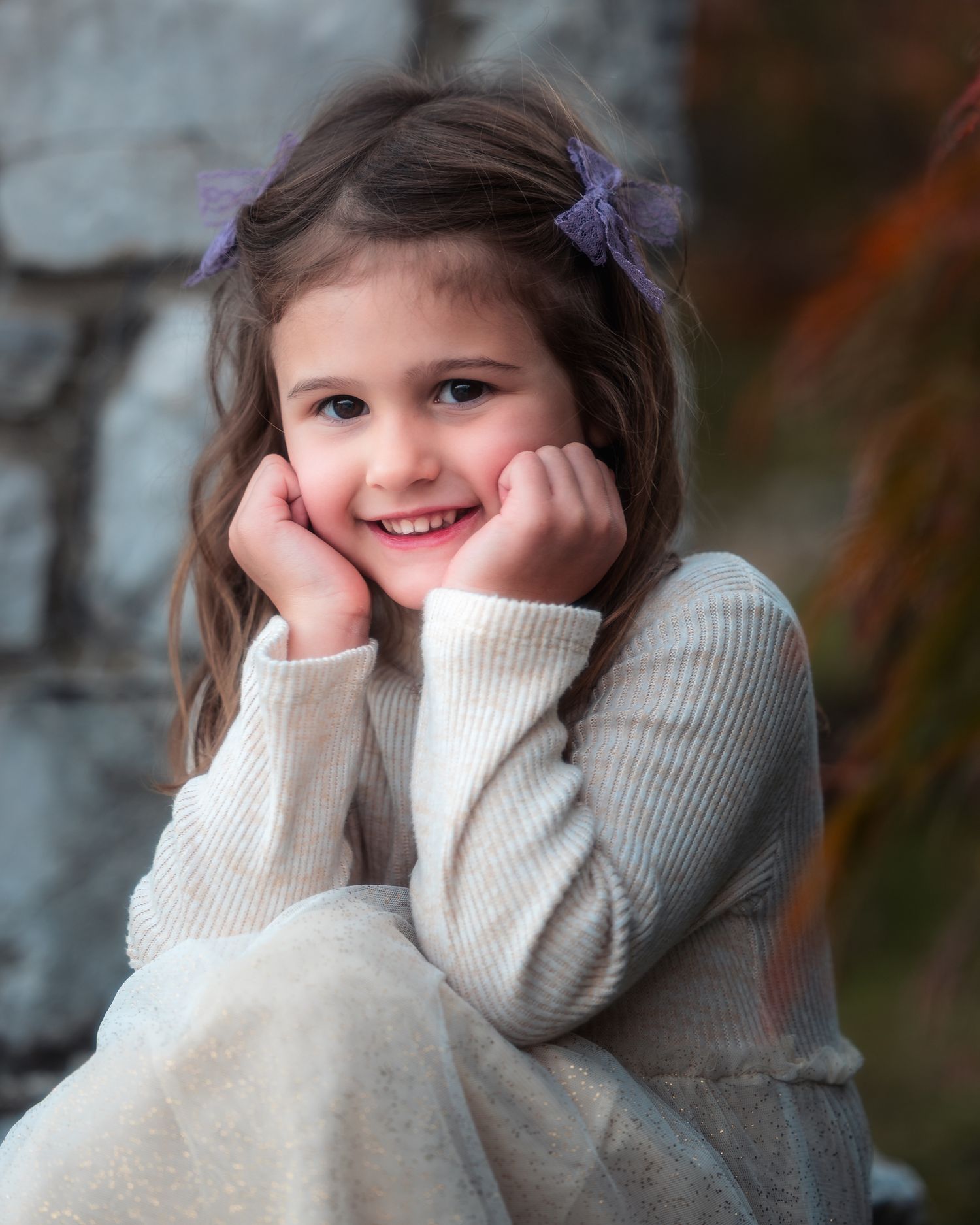 A young child in a cozy cream sweater and purple hair accessories poses with hands resting on cheeks.