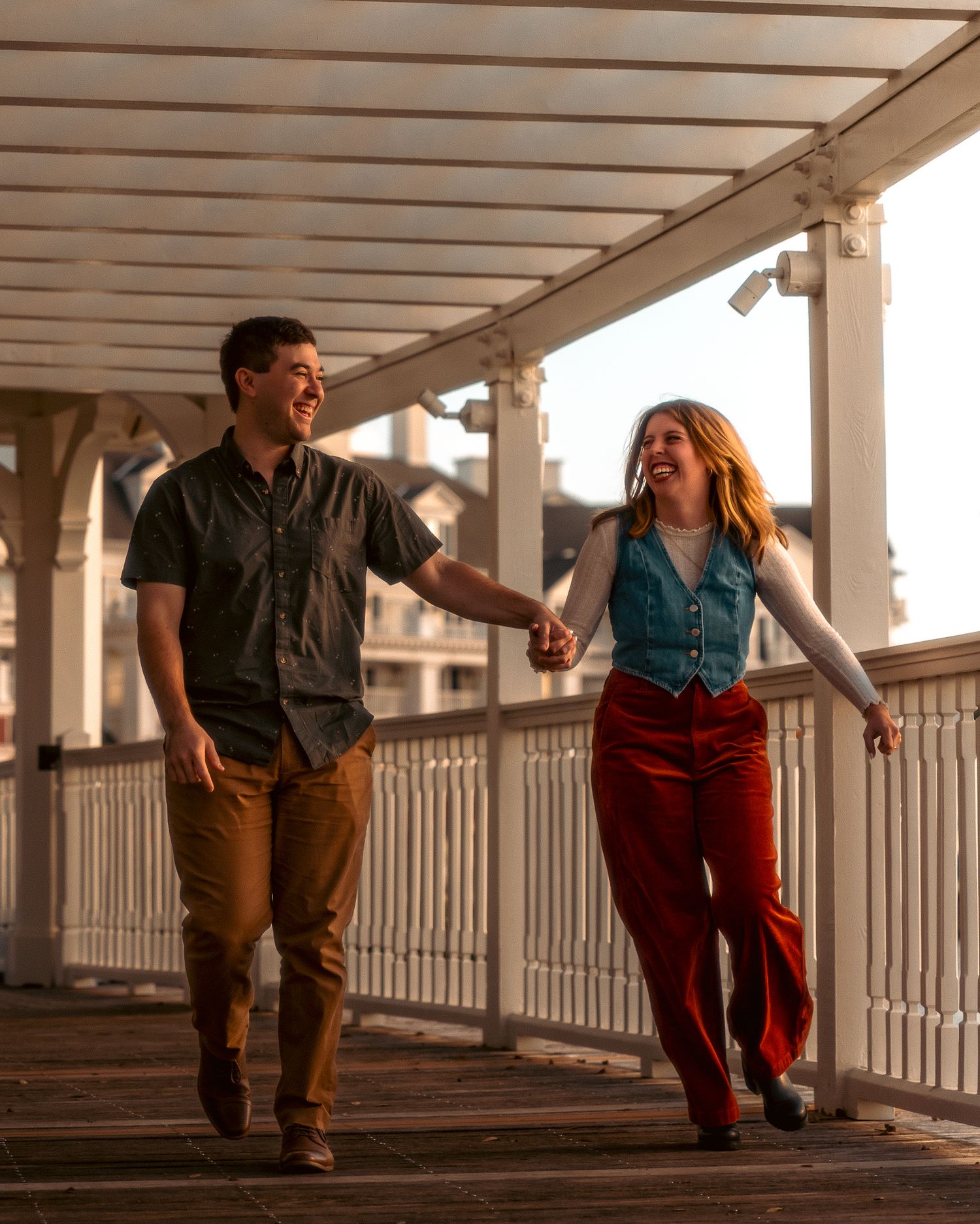 A couple holds hands while walking along a covered porch at sunset, wearing casual attire and smiling joyfully.