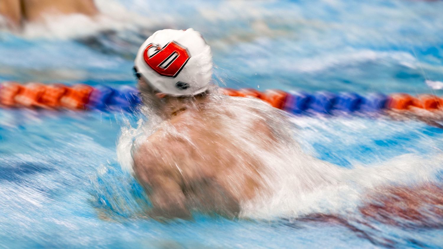 Competitive swimmer racing through blue pool water during breaststroke competition.