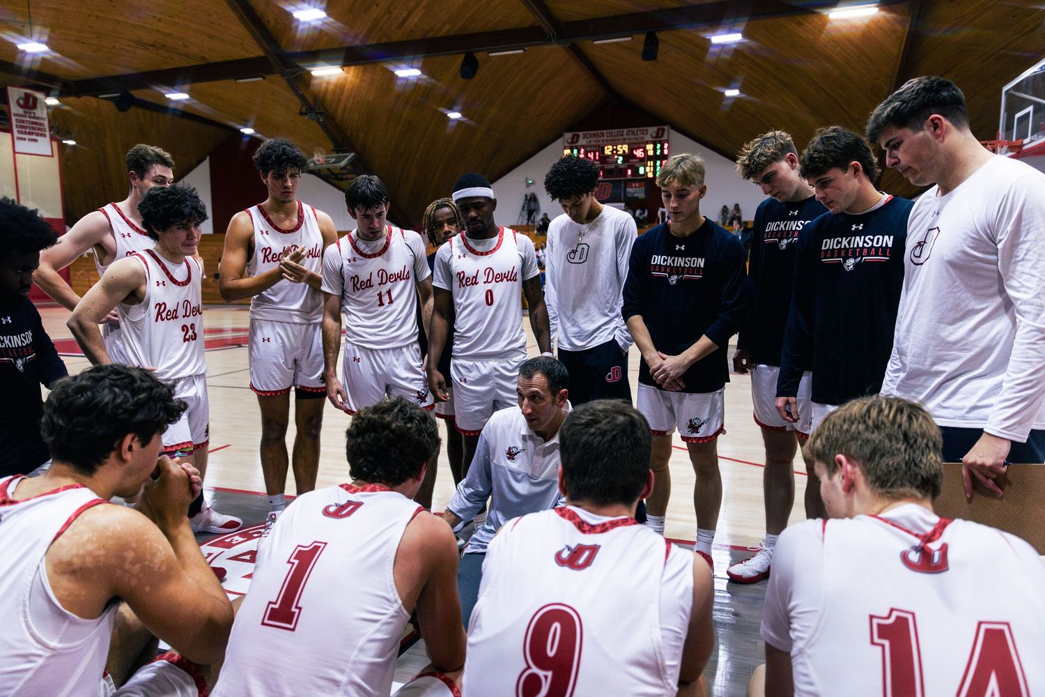 Basketball team huddles together in white jerseys during a game break in a brightly lit gymnasium.