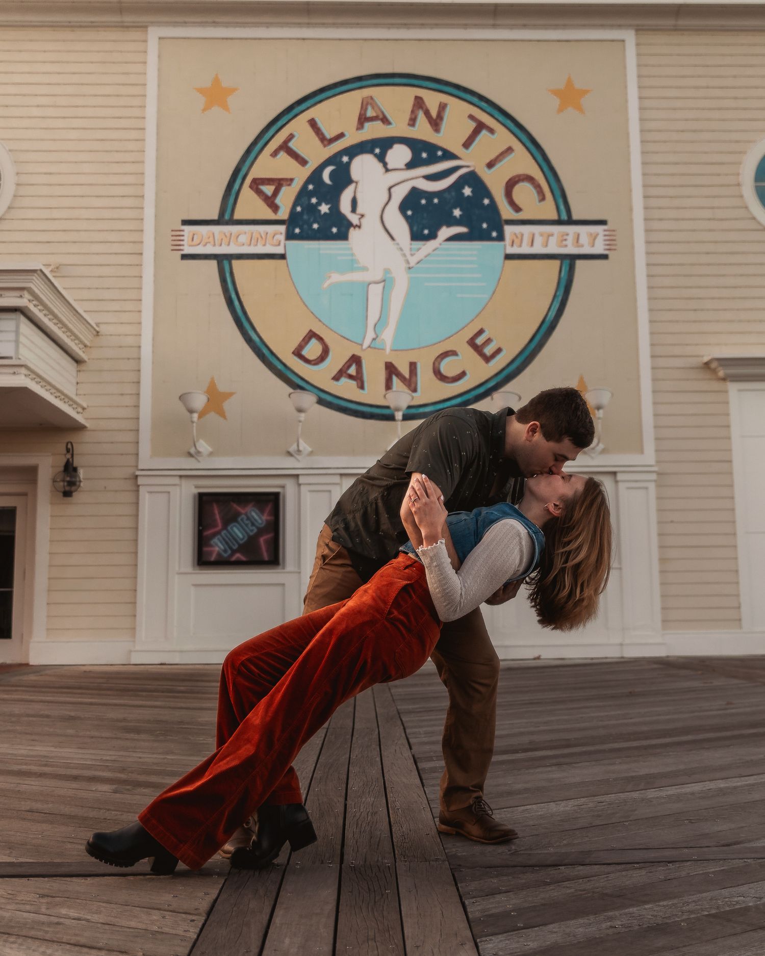A couple performs a romantic dance dip pose in front of the Atlantic Dance logo on a wooden boardwalk.