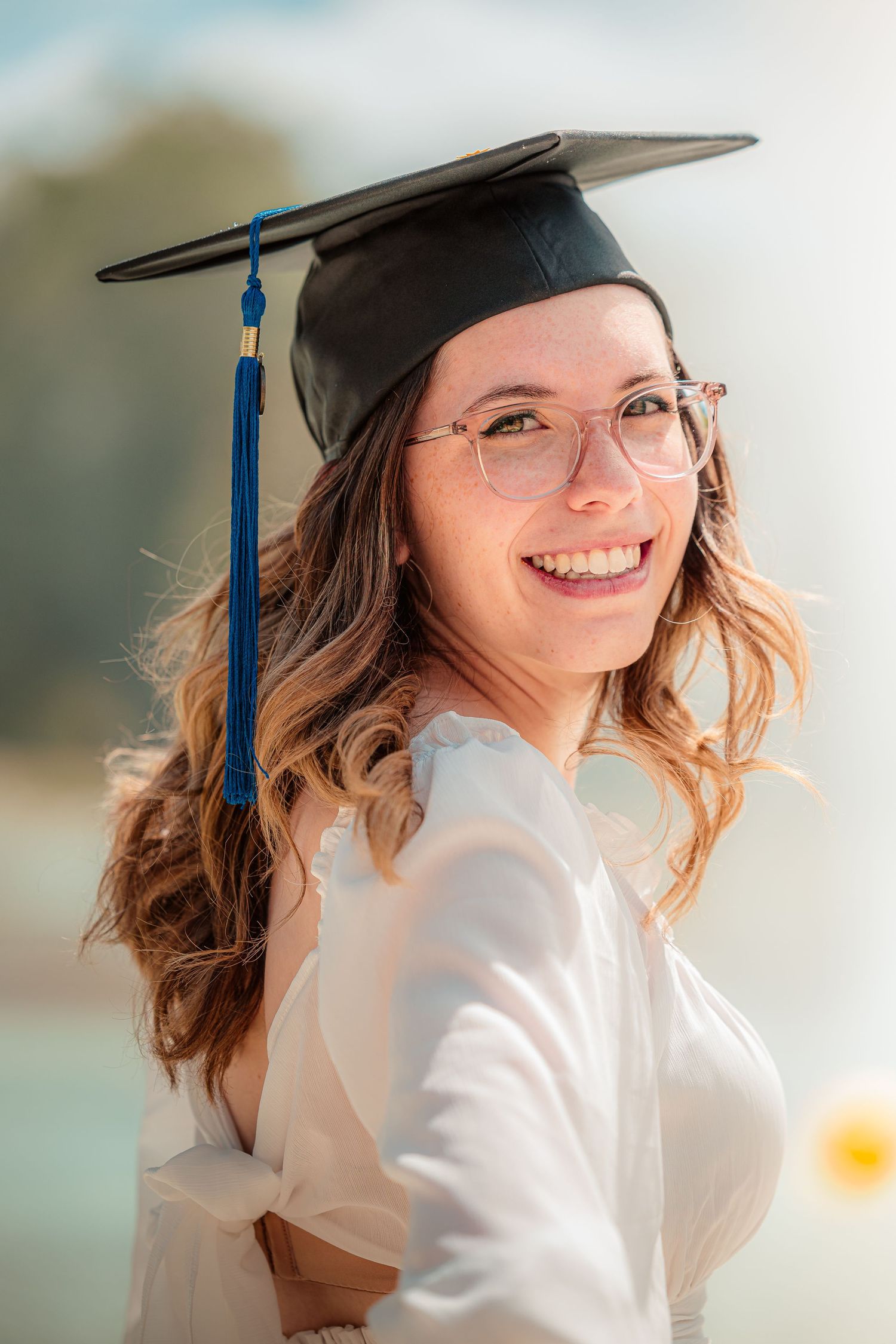 Graduate in cap and gown smiling while posing for graduation photos outdoors on a sunny day.