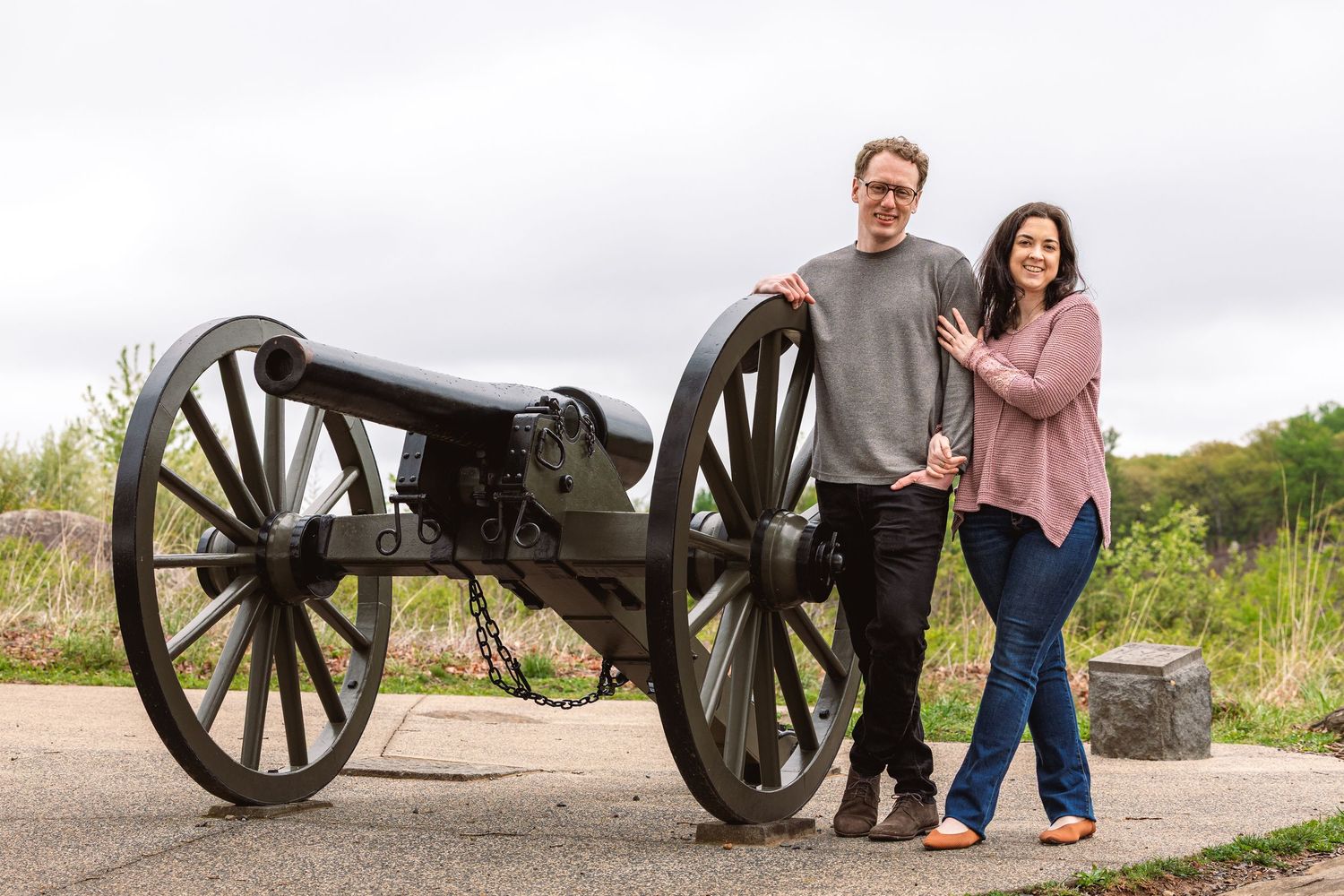 Couple posing together beside a historic Civil War cannon on a grassy battlefield under overcast skies. In Gettysburg National Military Park