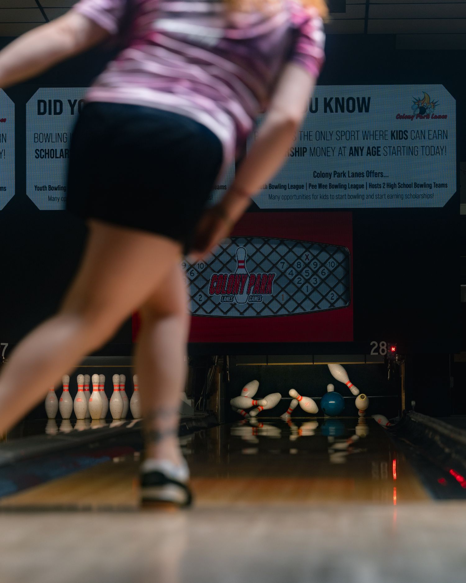A bowler releases a ball down a lane, scattering pins at a bowling alley with motivational signage in the background.