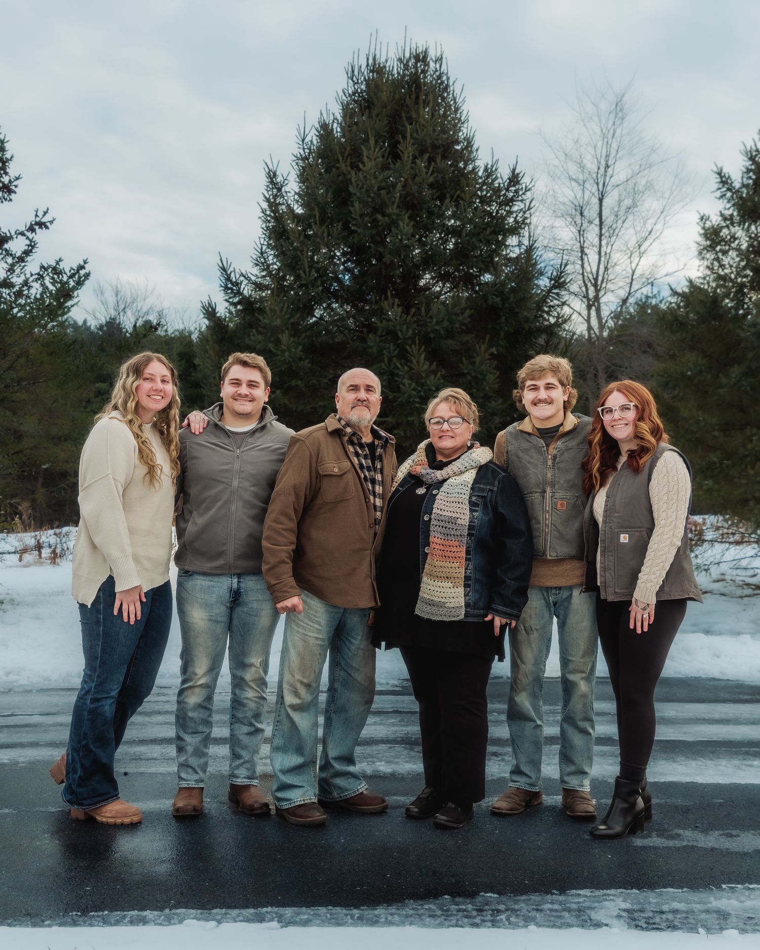 A group stands together outdoors in winter on a snowy paved surface with evergreen trees in the background.