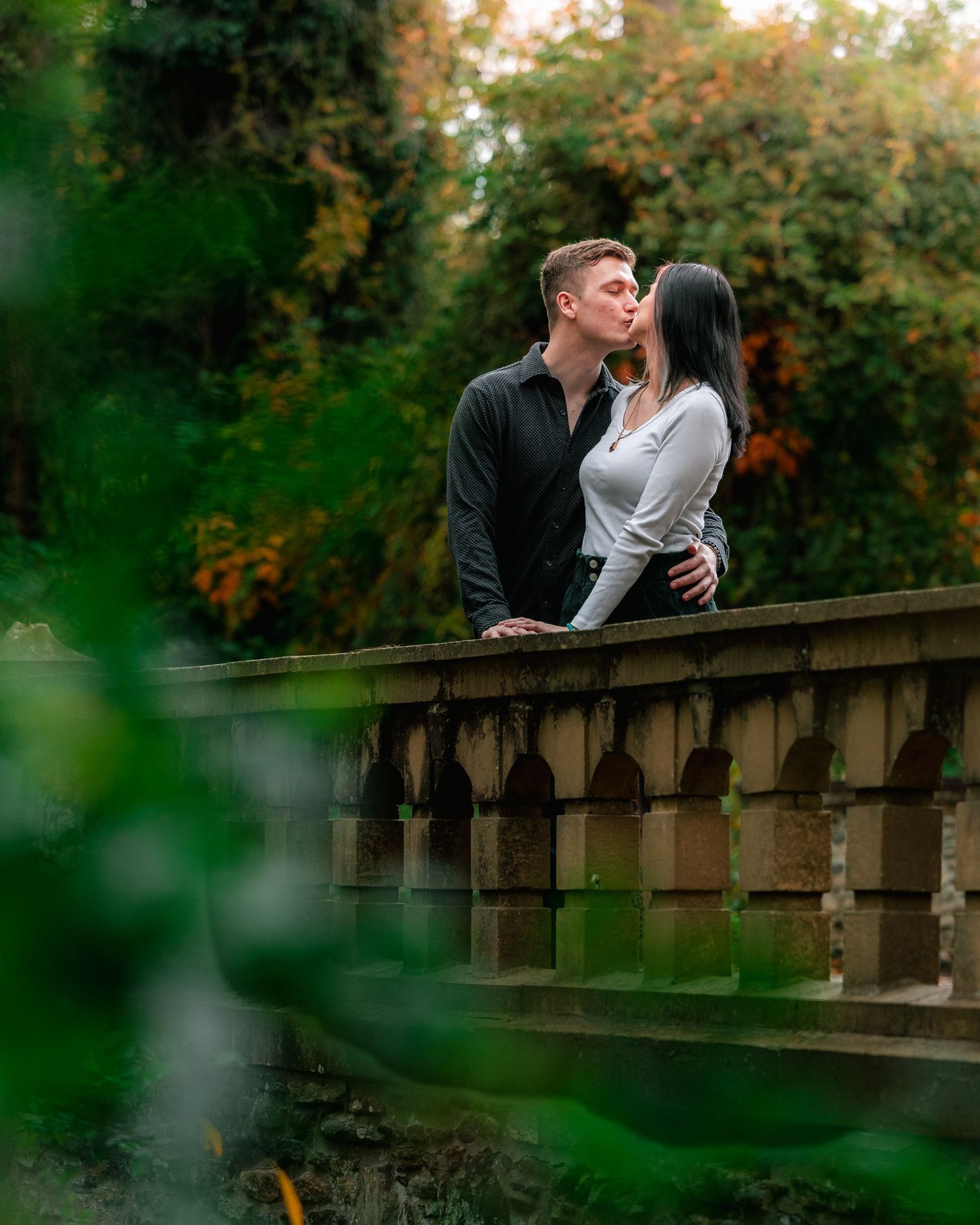 A romantic couple embraces on a stone bridge surrounded by autumn foliage and green leaves in the foreground.