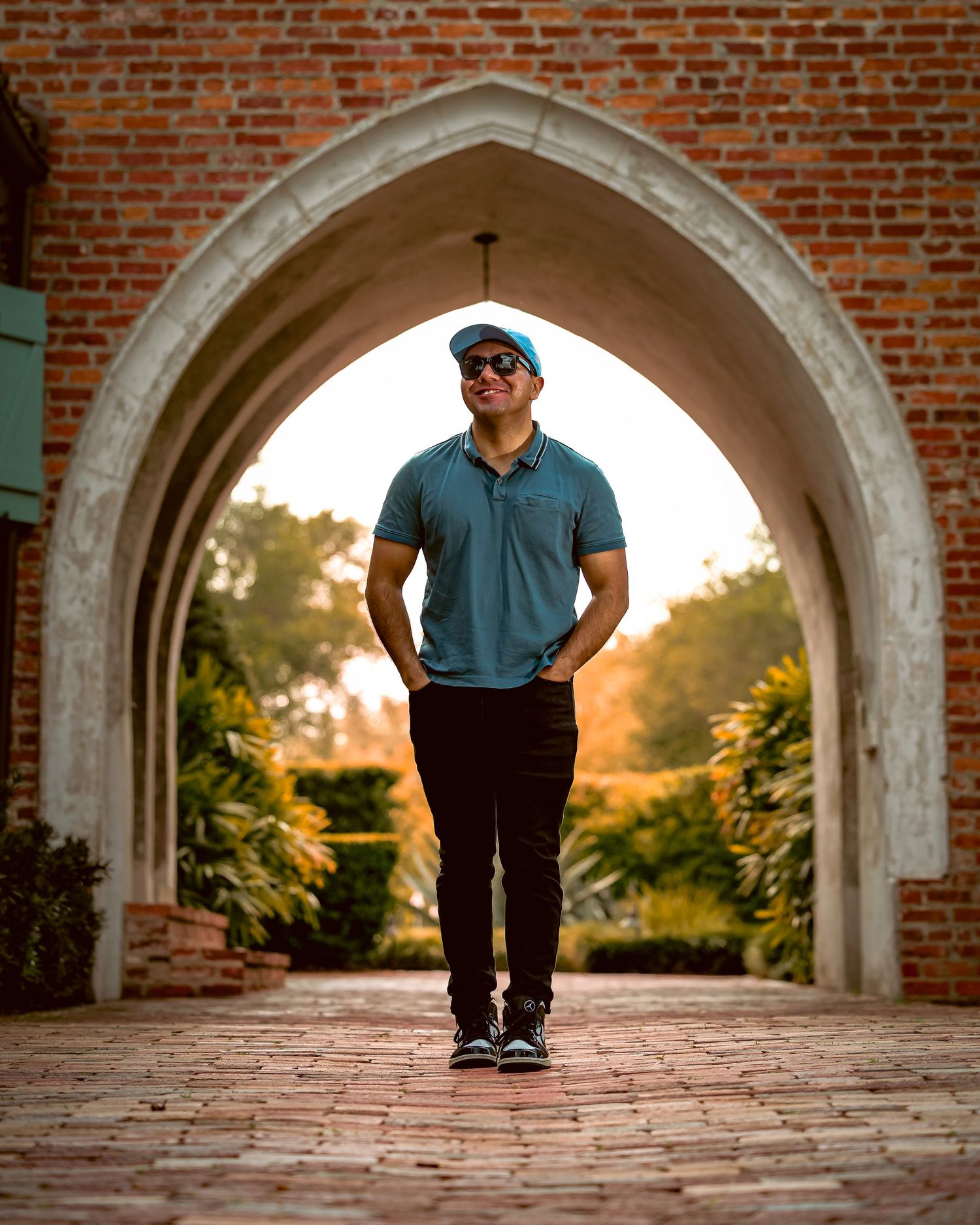 Someone in a blue polo and sunglasses stands under a brick archway at sunset with garden scenery in the background.