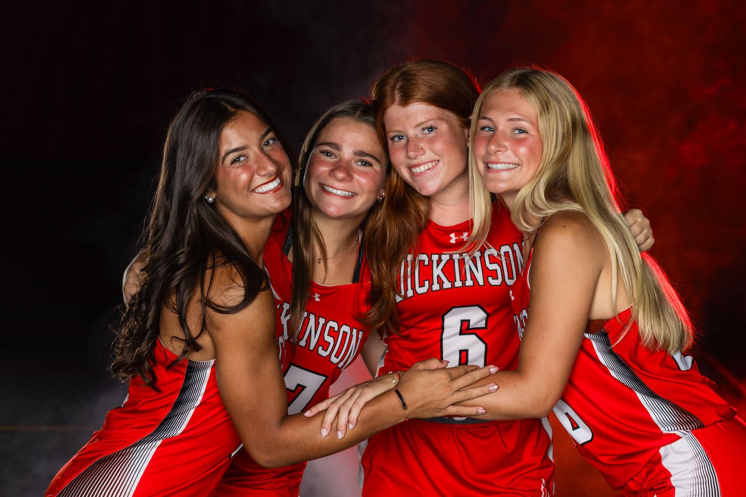 Four smiling girls in red Dickinson jerseys pose together for a team photo against a dramatic backdrop.