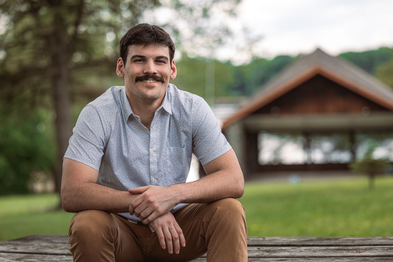 A person in a gray polo shirt sits on a wooden bench outdoors with a rustic building in the background.