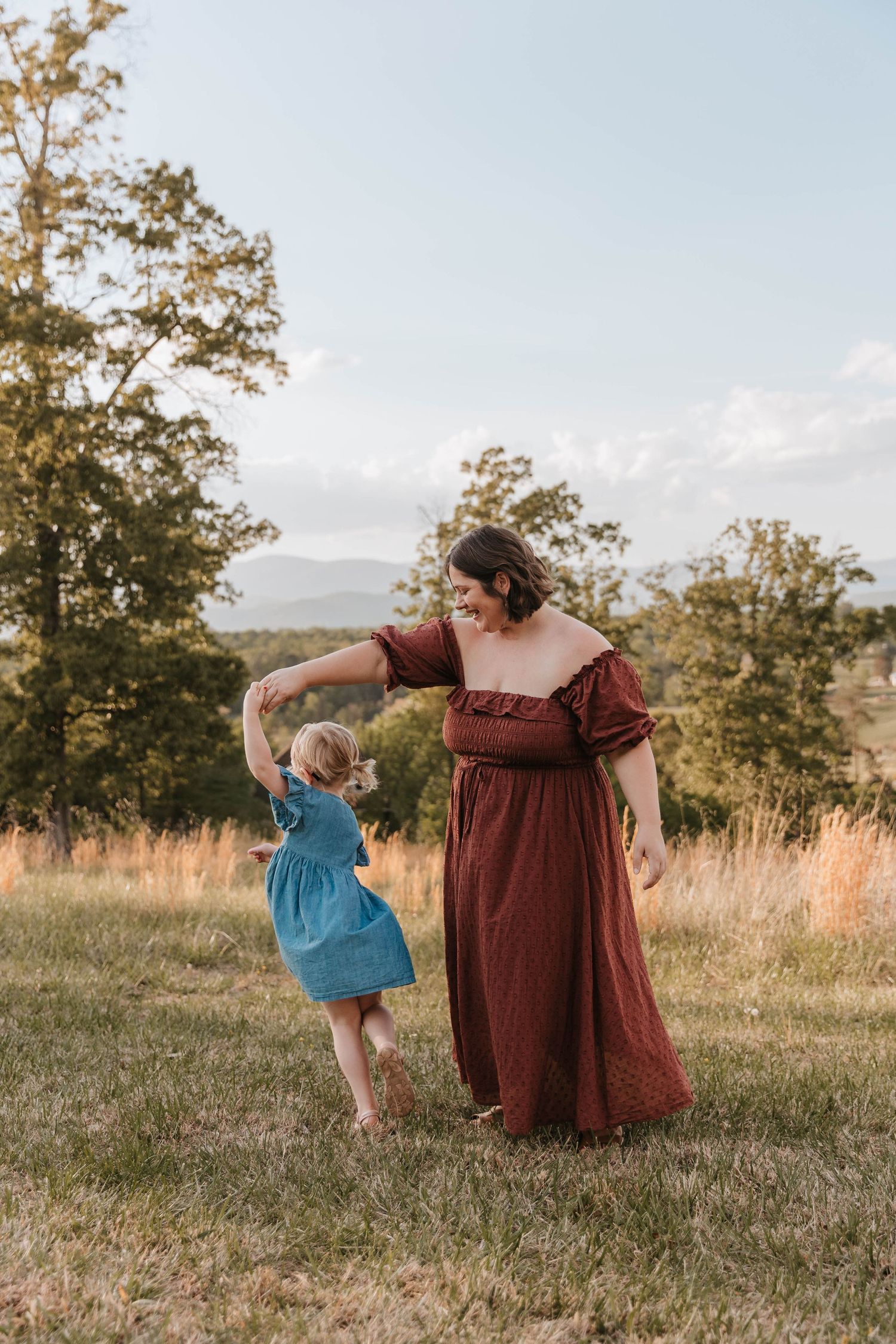 A woman in a rust dress twirls and dances joyfully with a little girl in a blue dress in a grassy mountain meadow.
