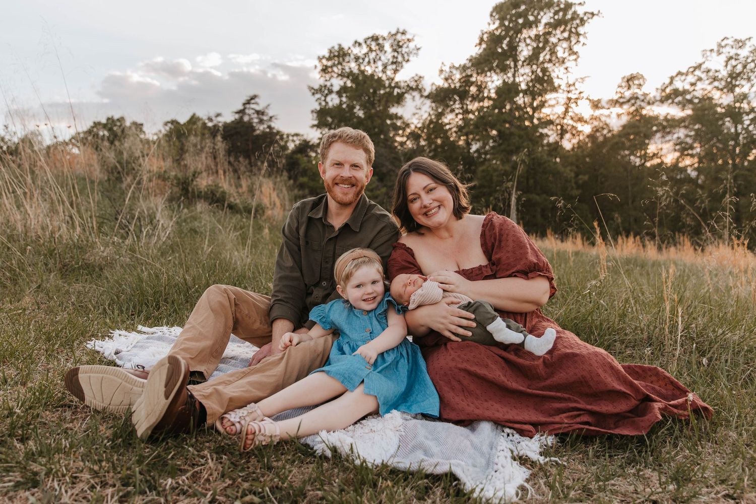 A family of four with a newborn, toddler girl, mom and dad sit together on a blanket in a grassy field at sunset.