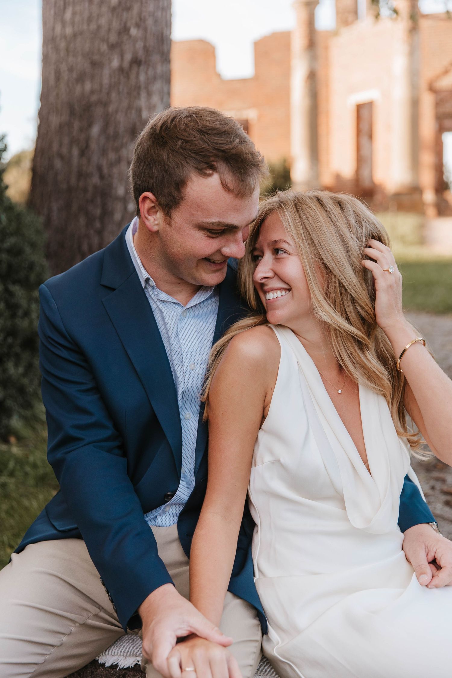 Smiling engaged couple sits close together outdoors, the man in a navy blazer and the woman in a white dress, with brick ruins behind.