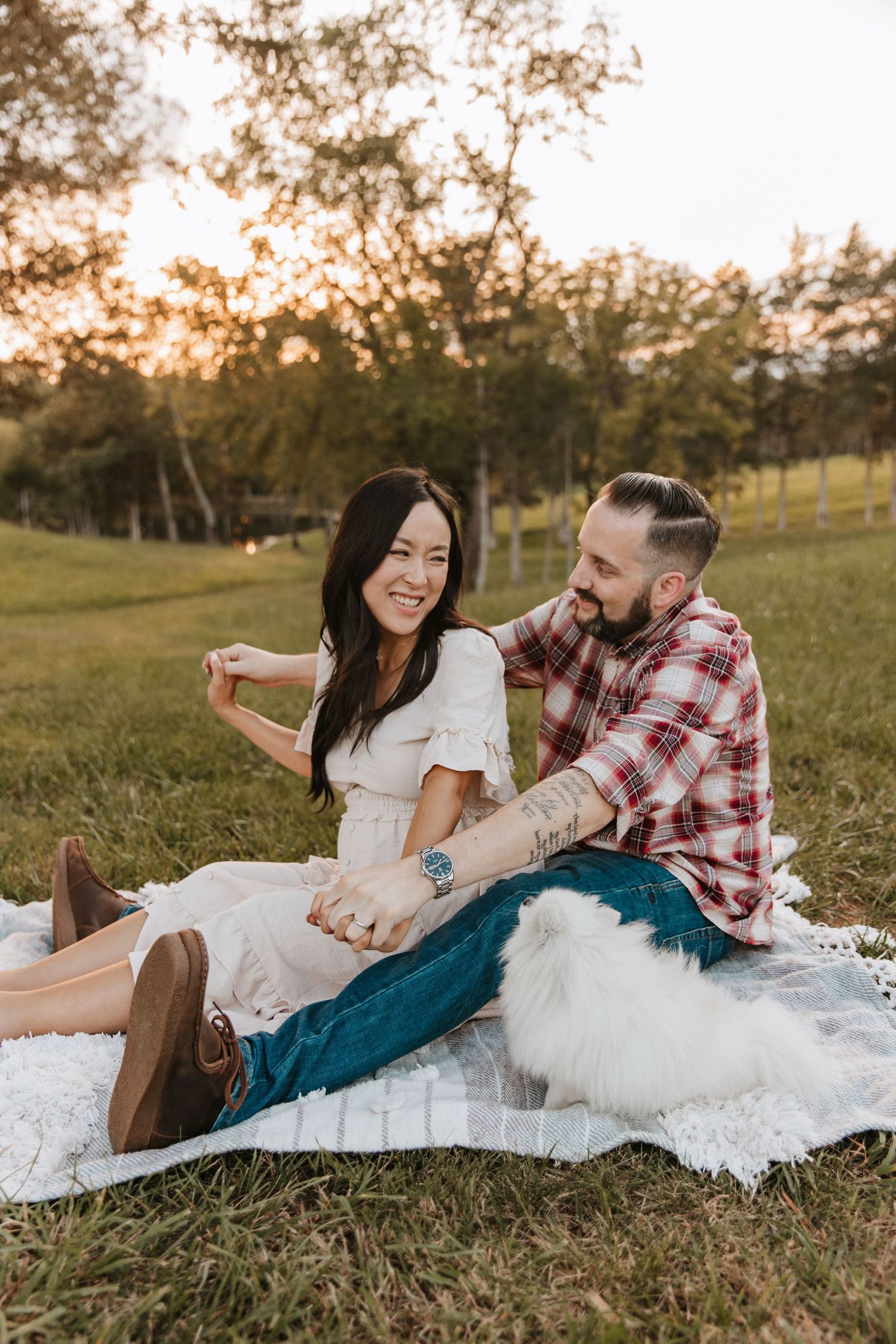 A laughing couple sits on a white blanket with a fluffy white dog as the sun sets behind trees in a pasture.
