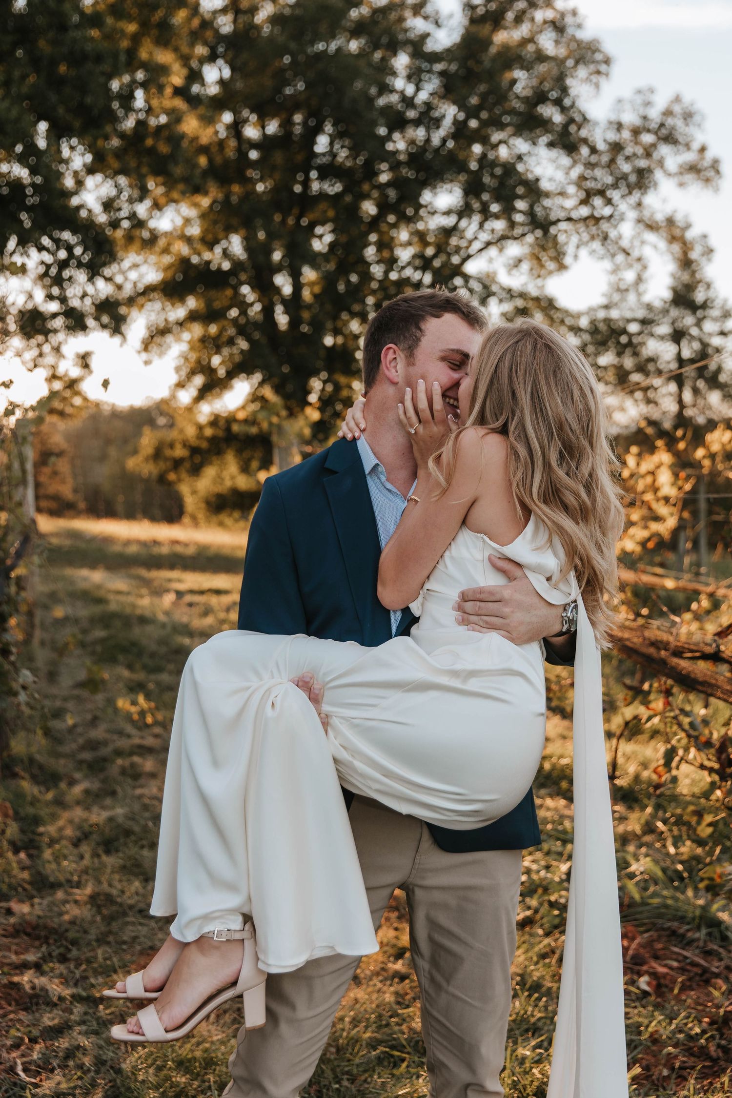 A man in a navy blazer lifts a woman in a flowing white dress into his arms for a kiss at golden hour outdoors.