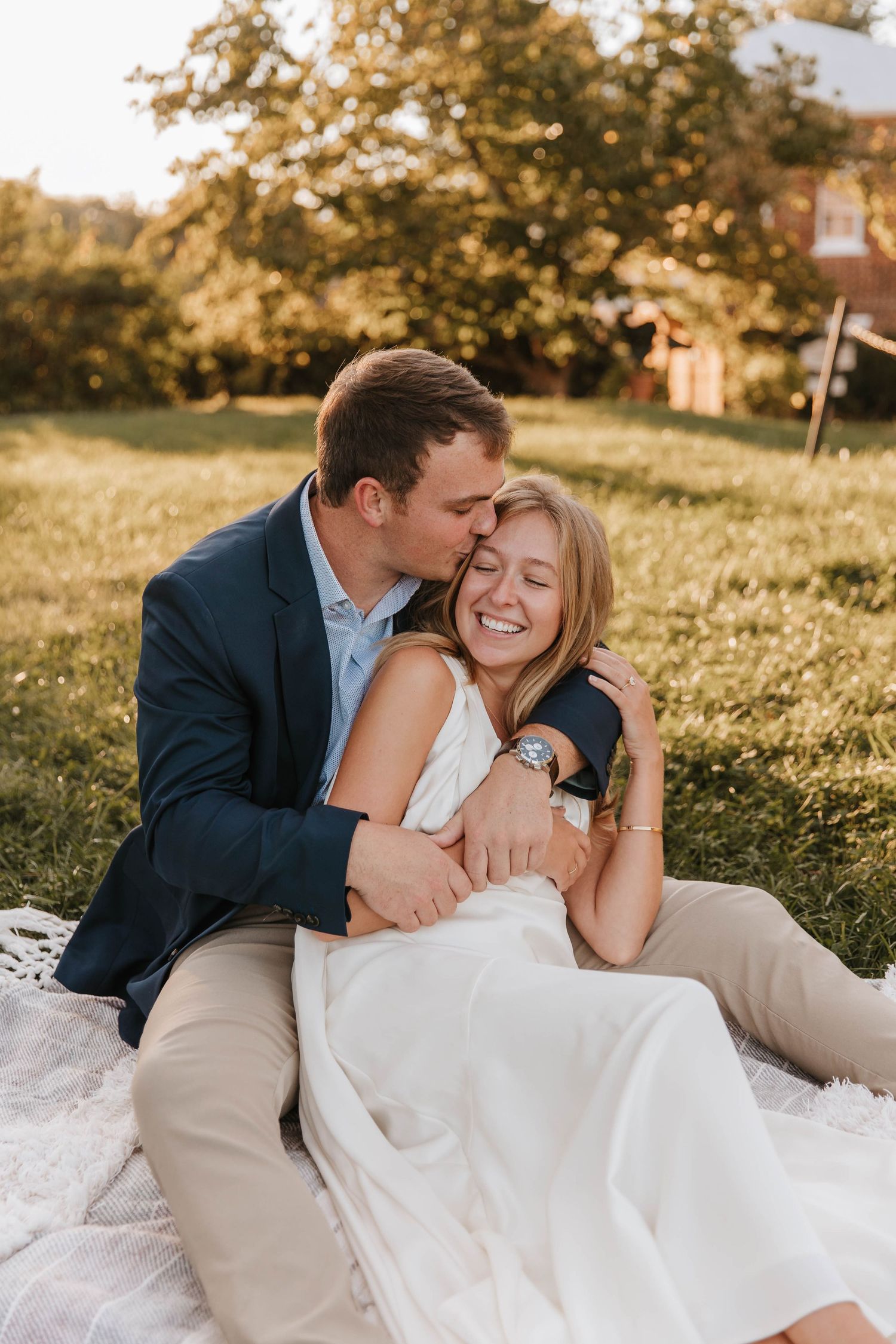 Man in a navy blazer kisses the forehead of his smiling fiancée in a white dress as they sit on a blanket in a sunny park.