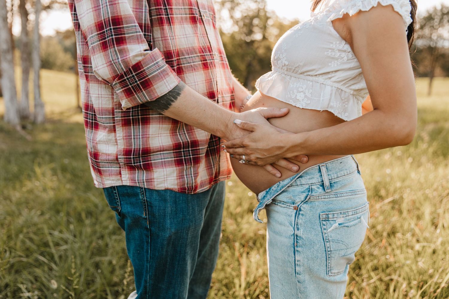 A couple in a plaid shirt and denim gently cradle a pregnant belly together in a sunny grassy field.