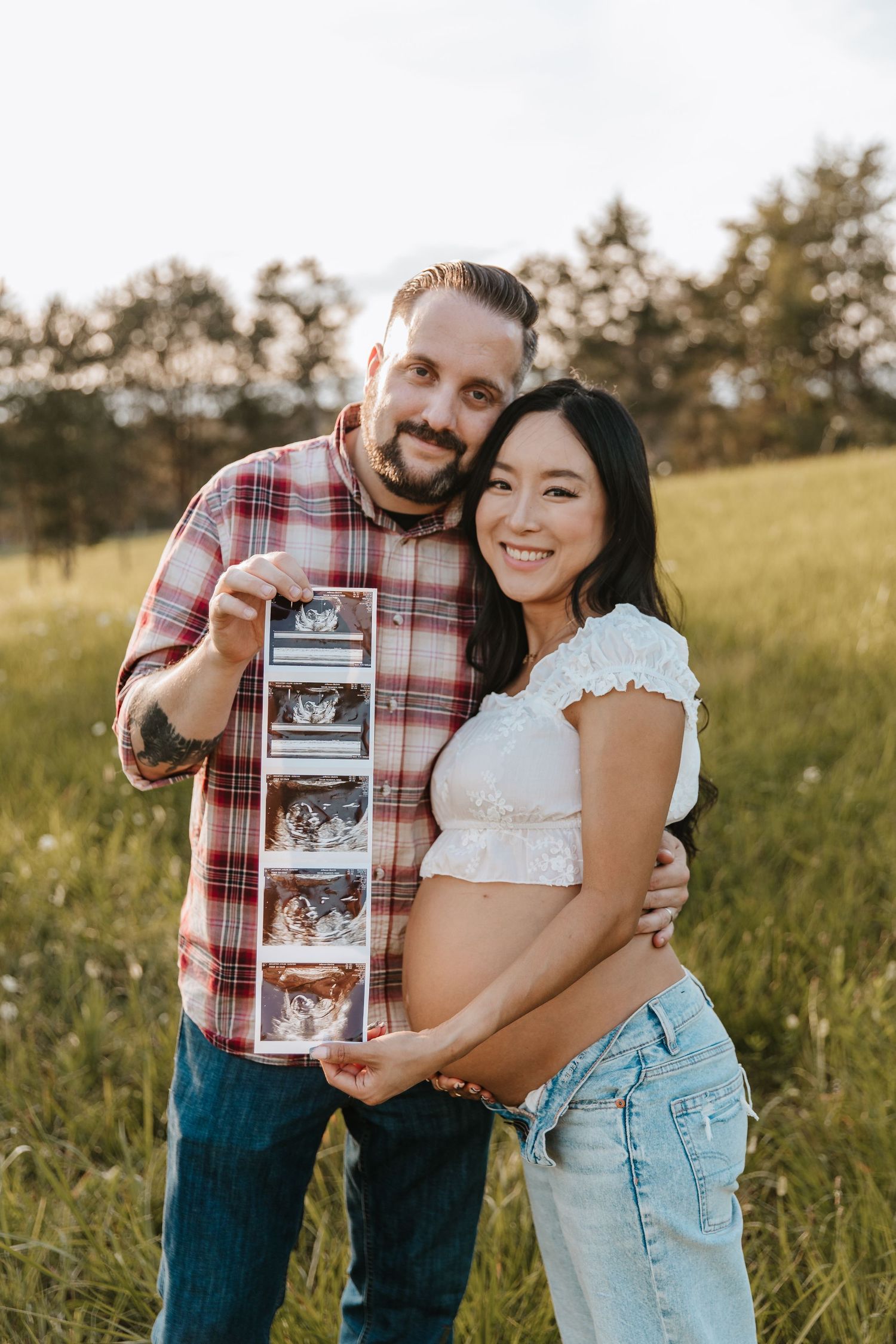 A smiling couple holds a strip of ultrasound photos while the pregnant woman cradles her belly in a green meadow.