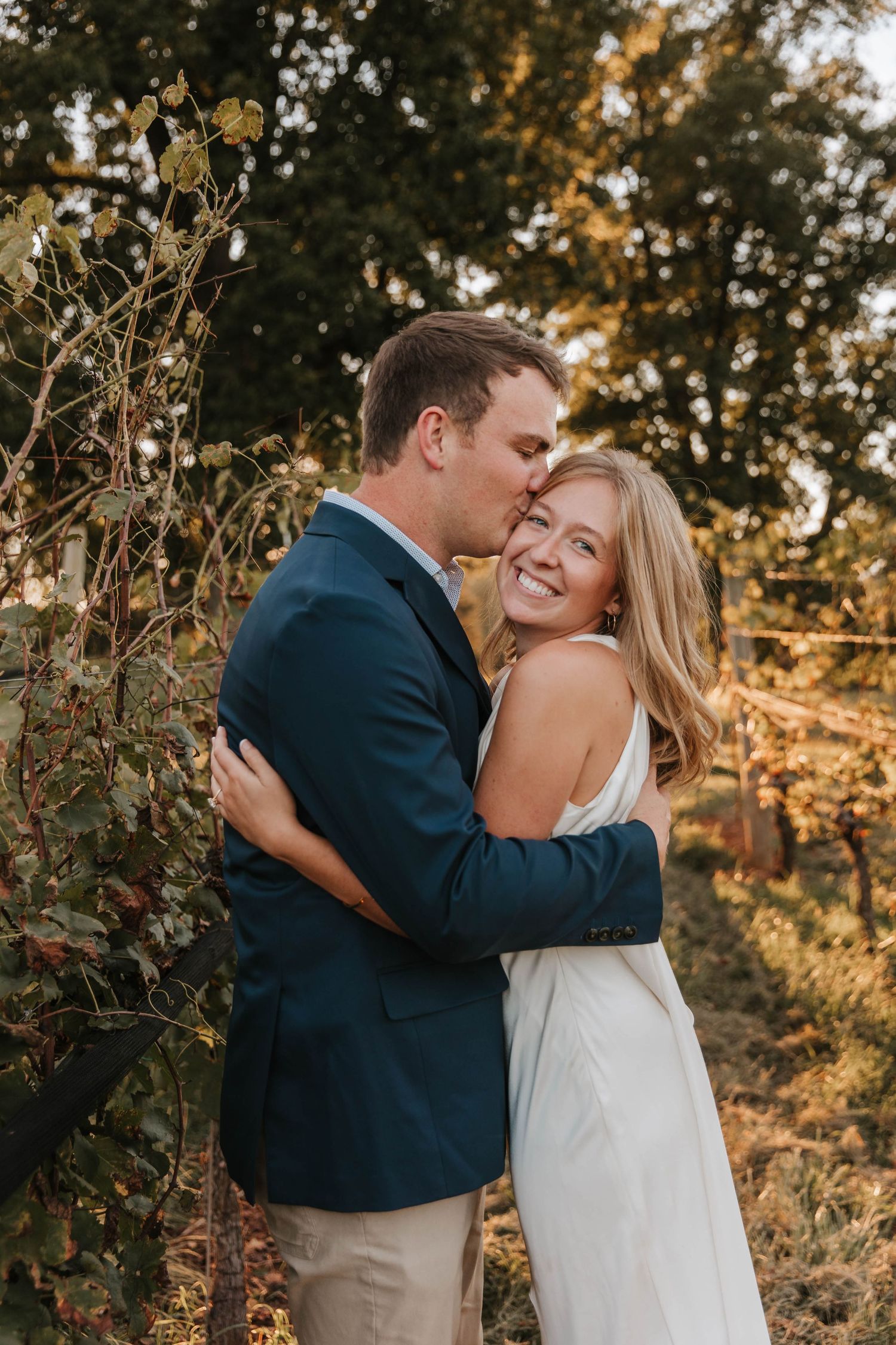 A smiling woman in a white dress embraces a man in a navy blazer as he kisses her cheek in a sunlit vineyard.