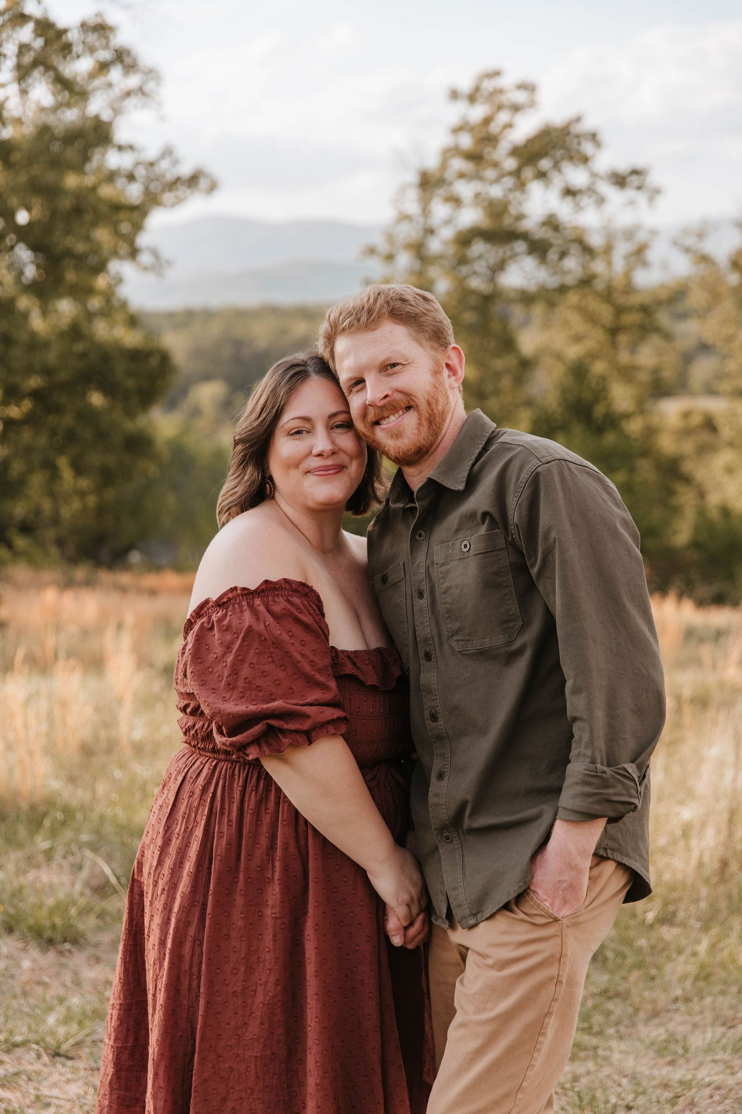 A smiling couple in a rust-colored dress and olive button-up stand close together in a field with mountains behind.