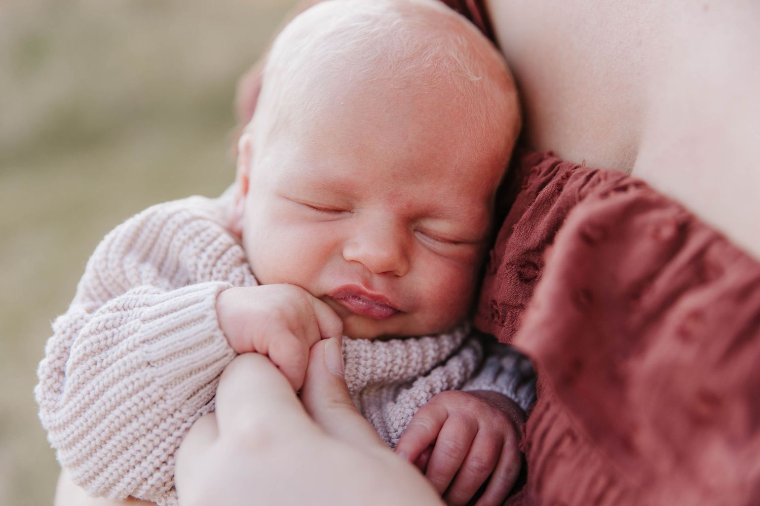 A sleeping newborn in a knit sweater rests peacefully in a mother's arms during an outdoor family photo session.