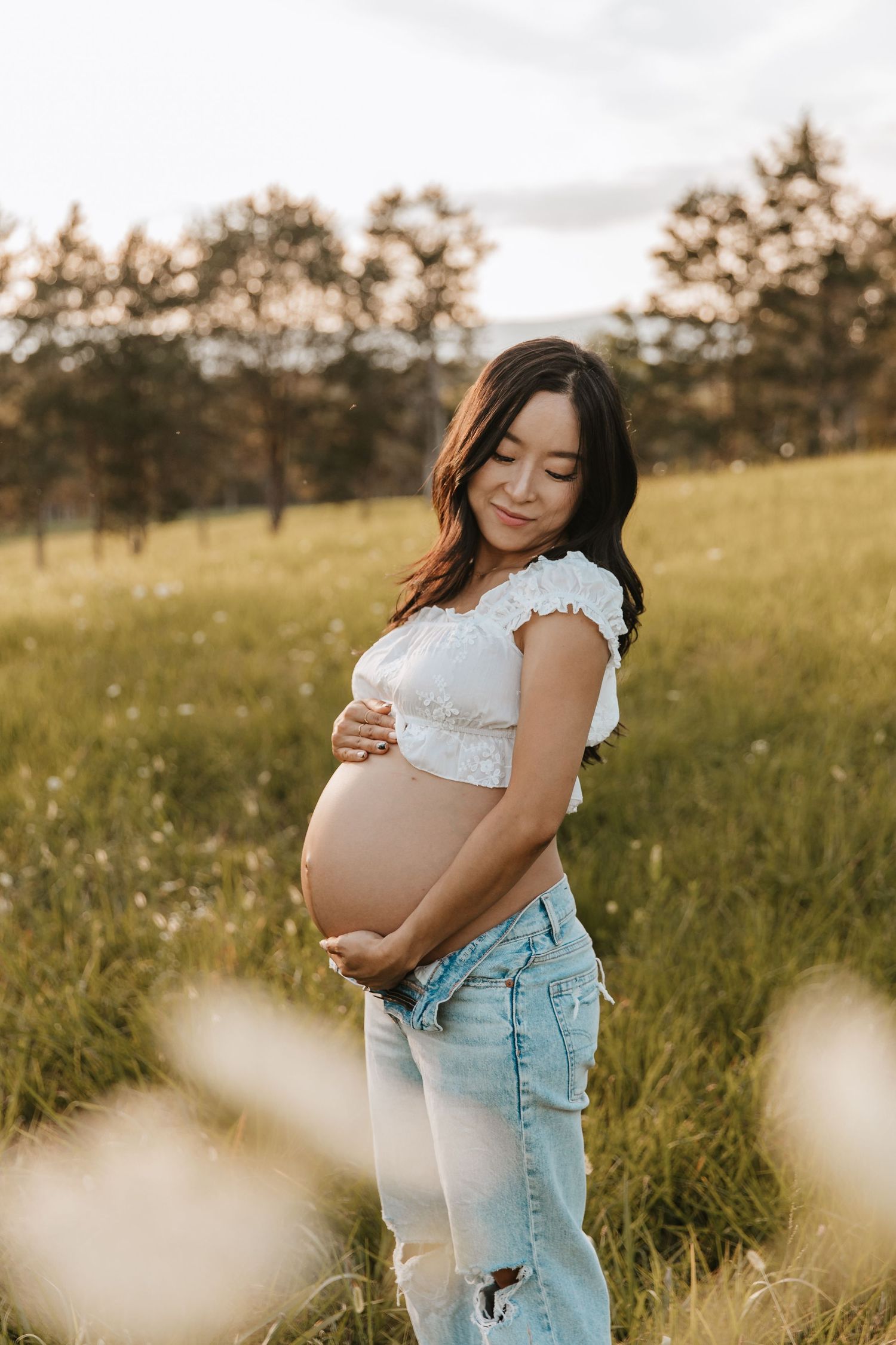 A pregnant woman in a white crop top and jeans smiles down at her bare belly in a golden grassy field.