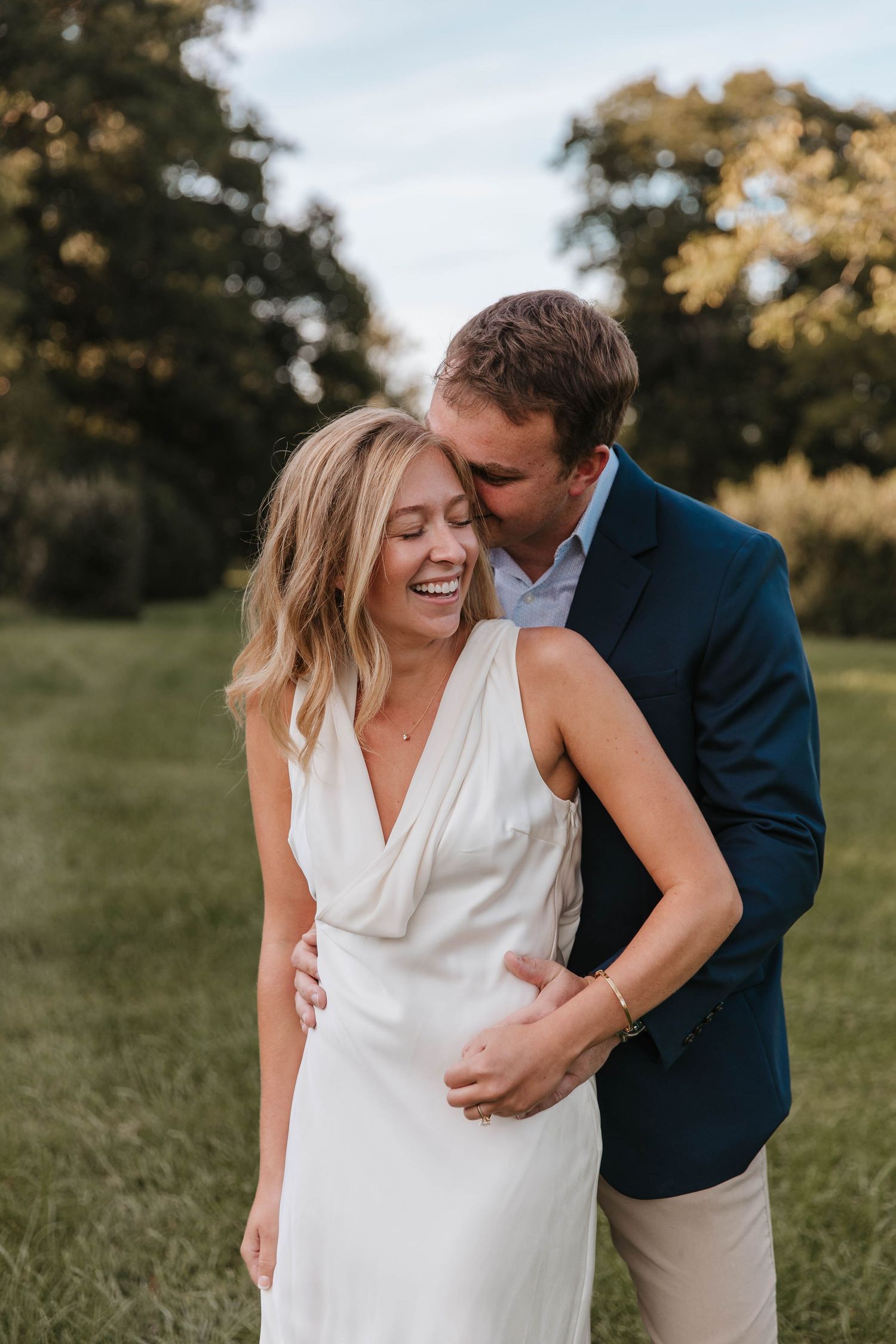 Laughing woman in a white dress is embraced from behind and kissed on the forehead by her fiancé in a navy blazer outdoors.