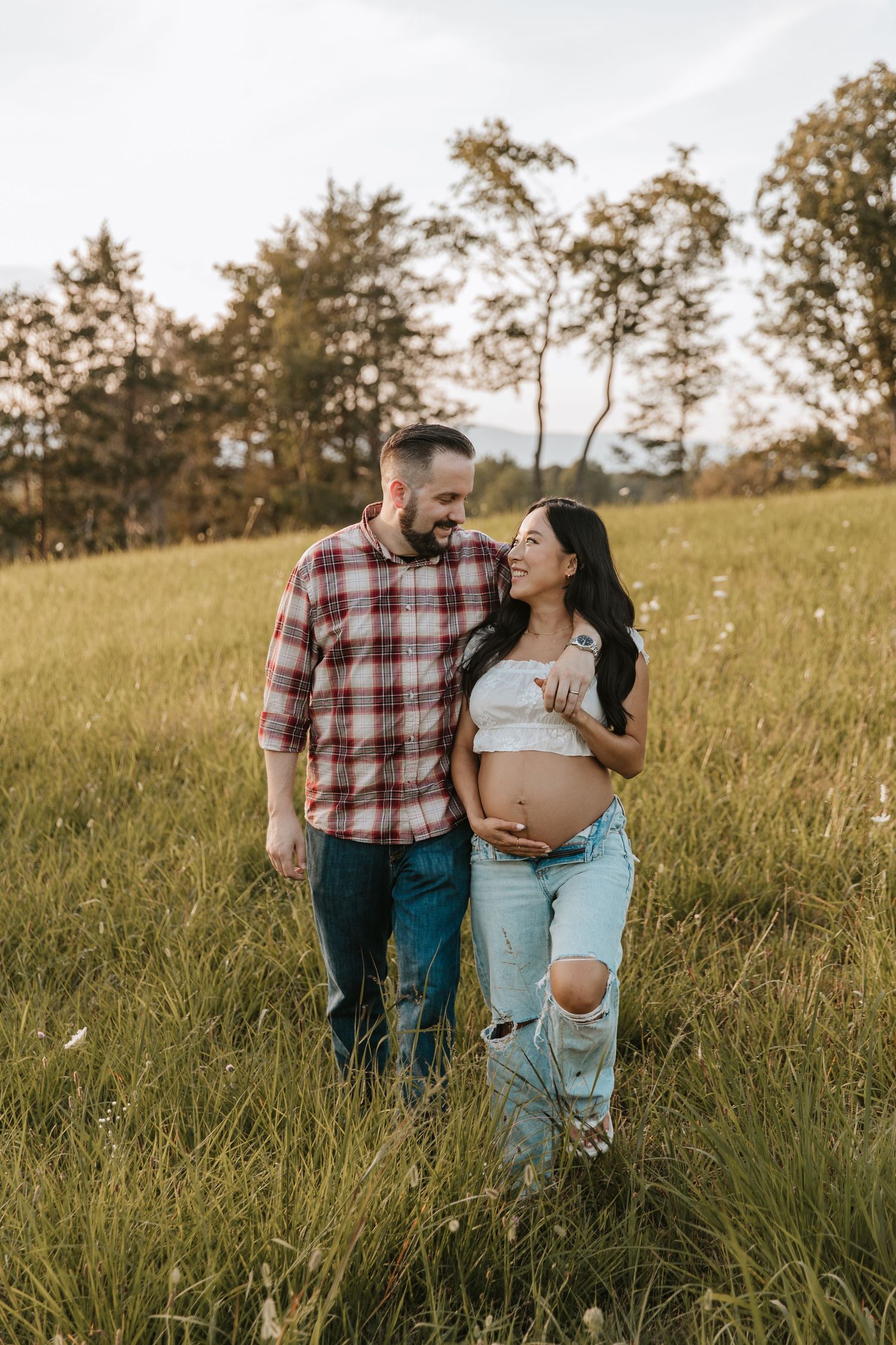 Expecting couple walks arm in arm through a green meadow as the smiling mom-to-be shows her baby bump in a cropped top.