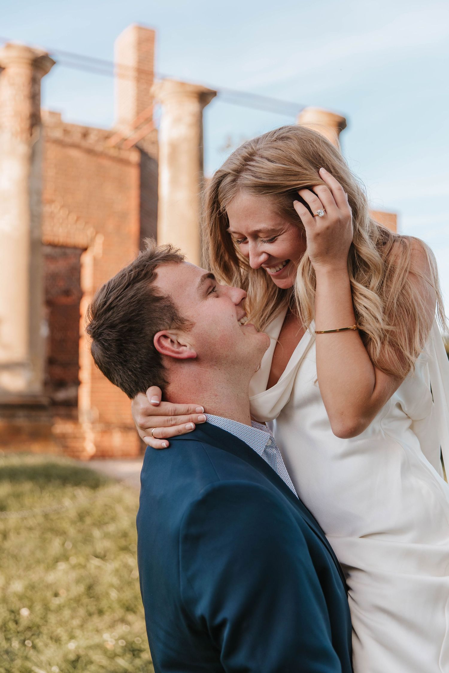 Joyful woman in a white dress smiles down at her fiancé in a navy blazer during an engagement session by historic brick ruins.