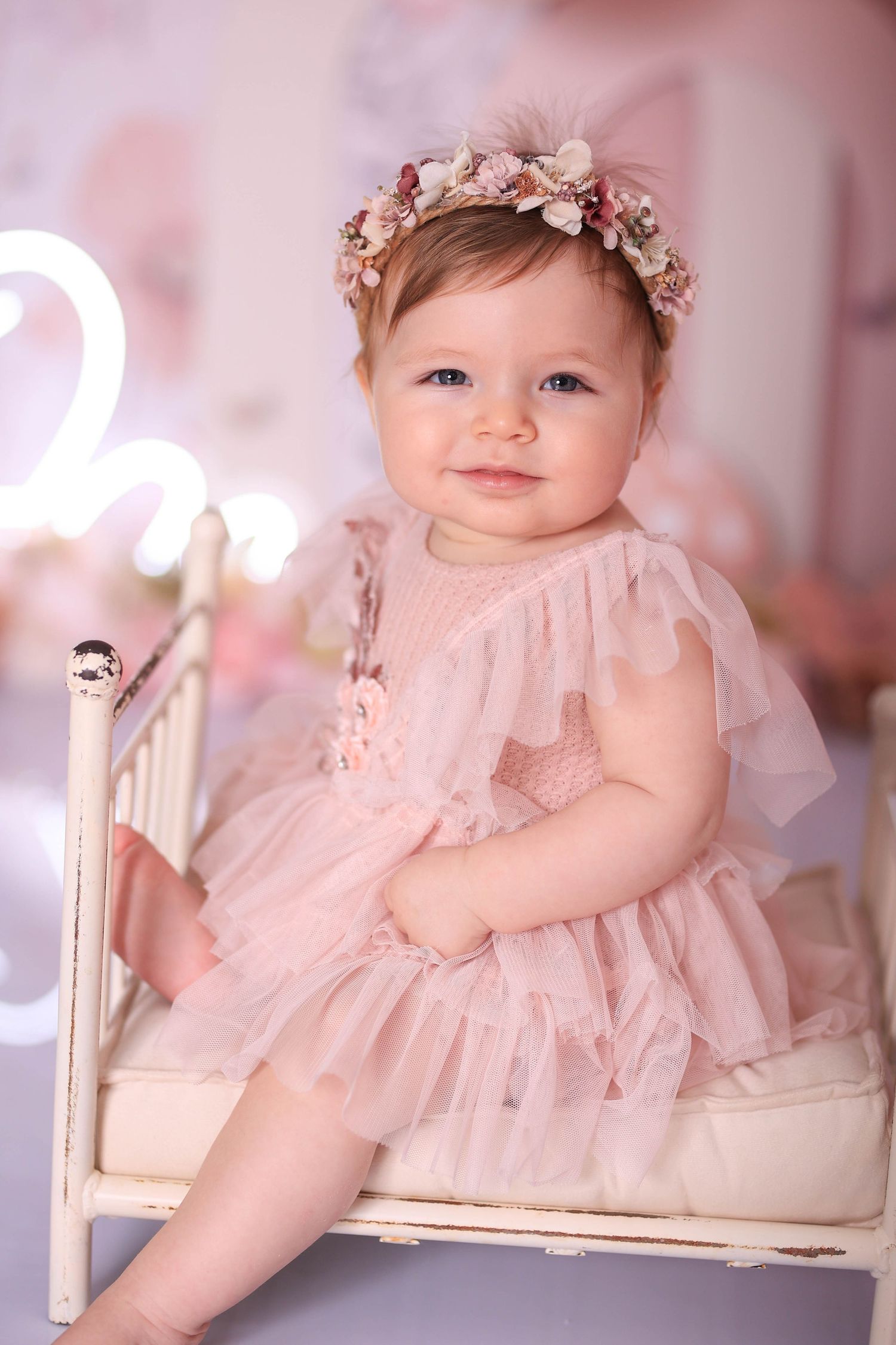 Baby in pink tulle dress and floral headband sits on white vintage chair against soft pink background. Dubai Photographer