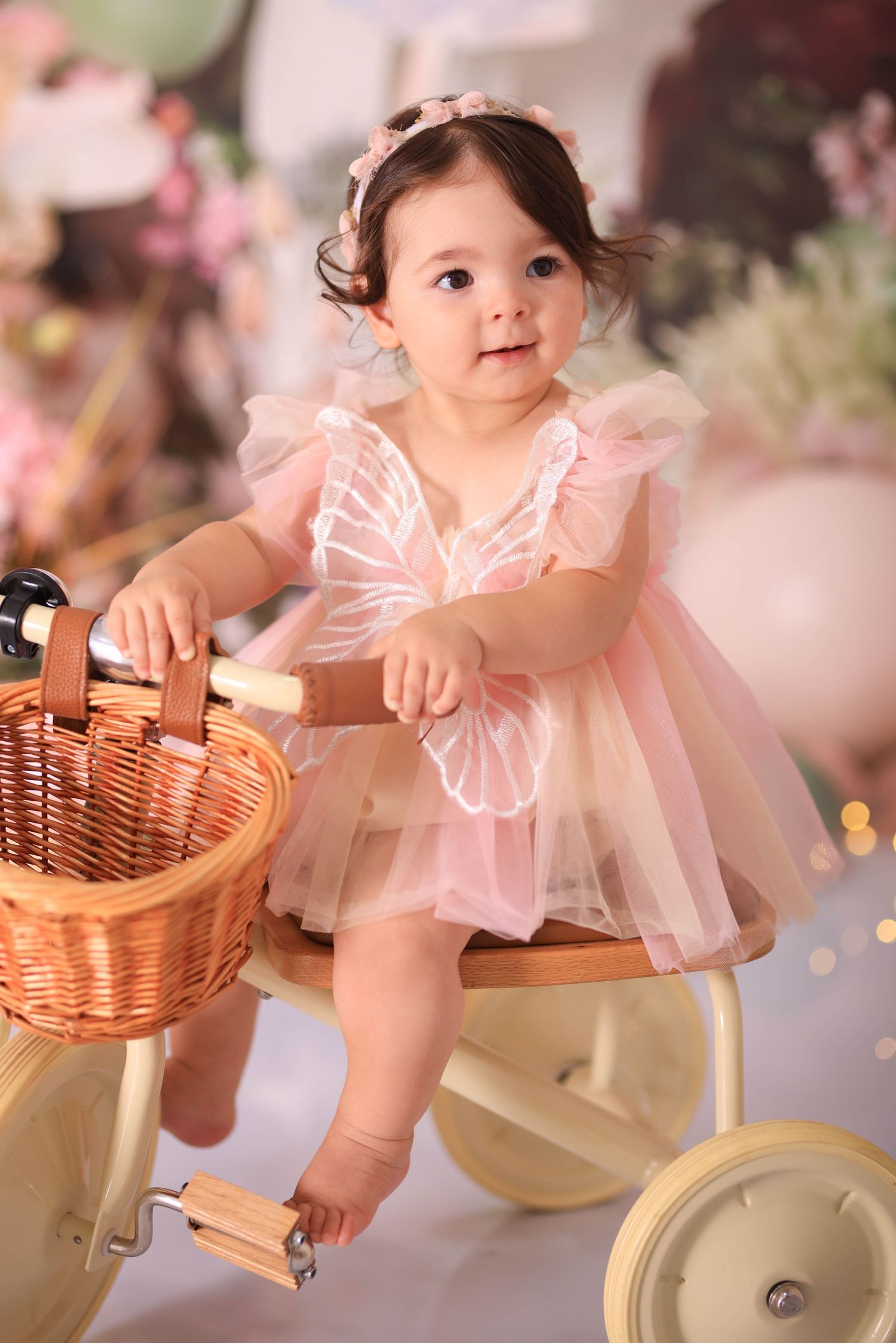 A small child in a pink tulle dress sits on a vintage bicycle with a wicker basket in a dreamy floral setting. Dubai Photographer