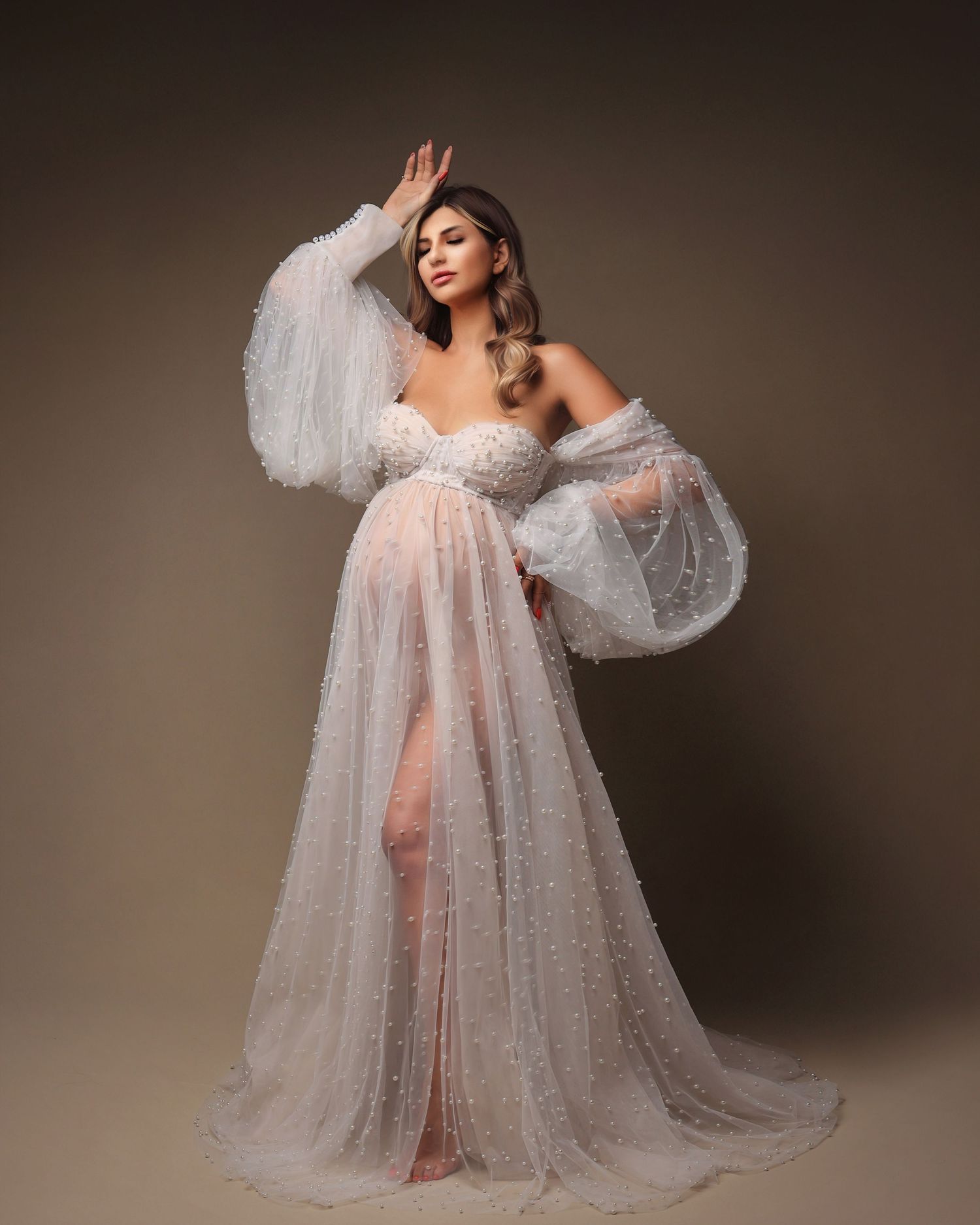 Elegant maternity portrait in flowing white tulle gown with dramatic sleeves against neutral background. Dubai Photographer